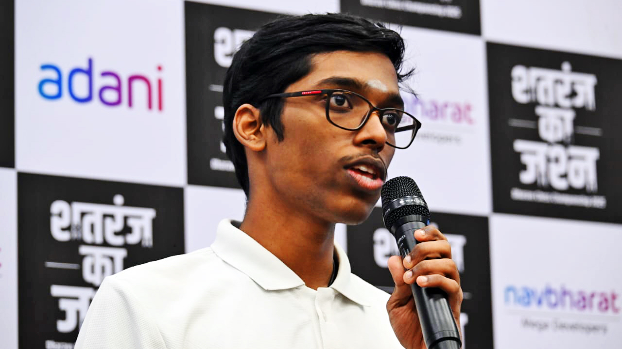Young man with glasses speaks into a microphone, "Adani" logo visible in background.