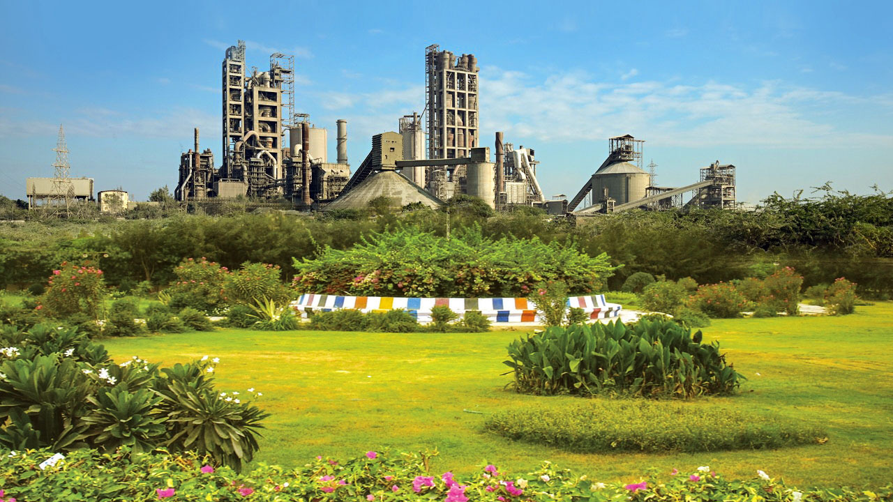 A sprawling industrial cement factory complex set behind a vibrant, well-kept green park with colorful borders.