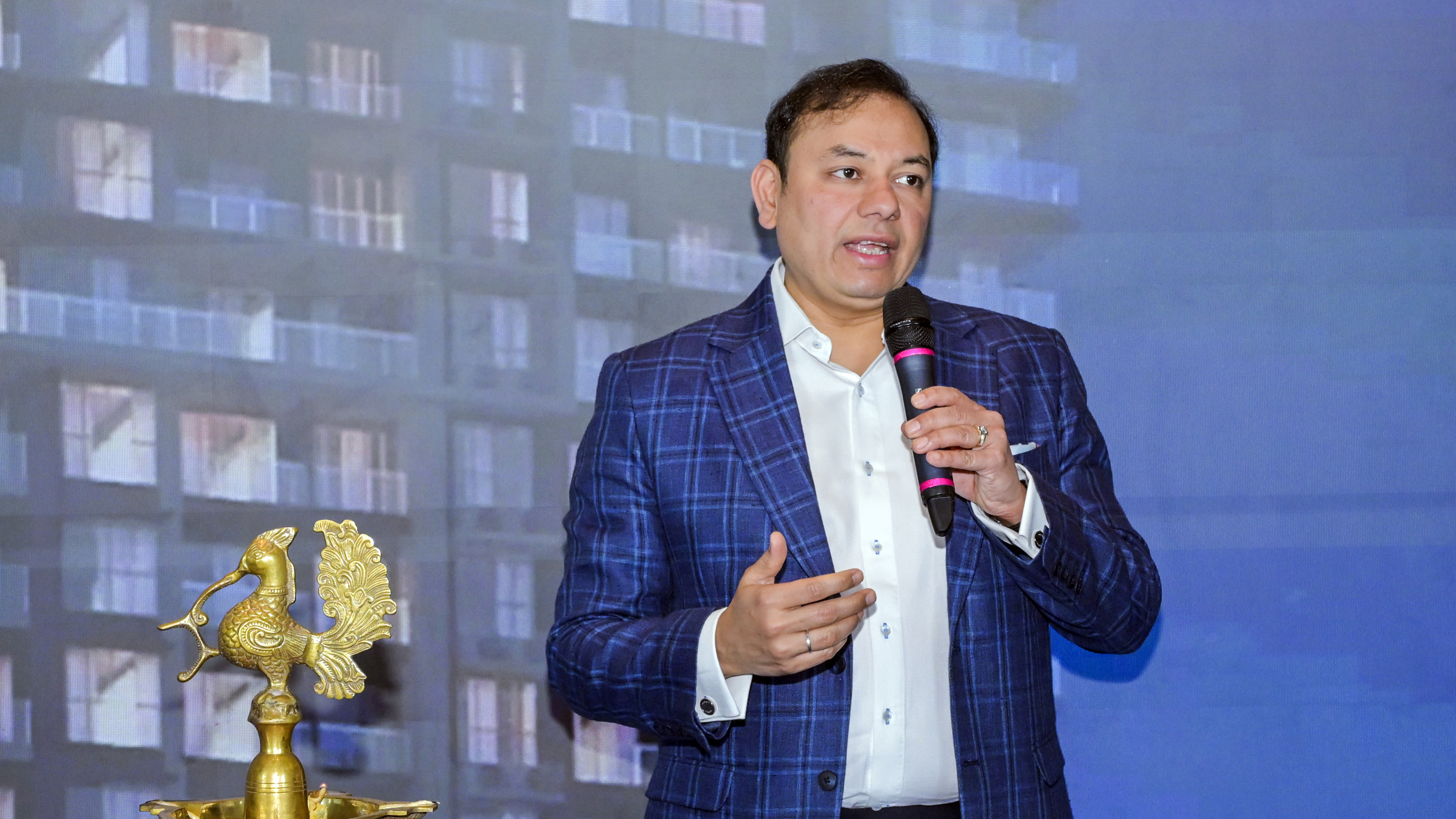 Man in blue plaid suit speaks into mic, with traditional brass peacock lamp and building background.