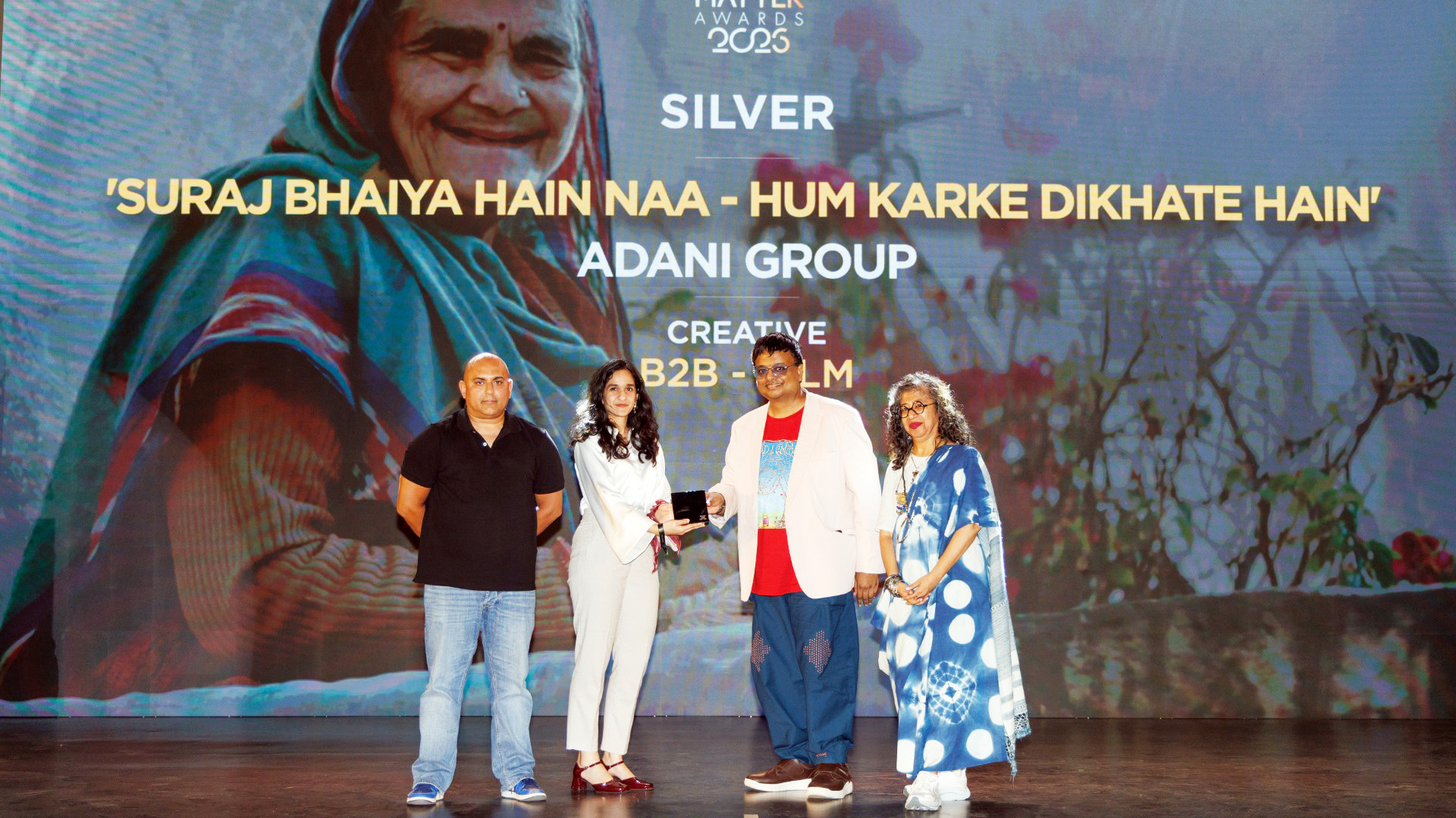 Four people on stage at an awards ceremony, receiving a Silver award from Adani Group, with a large screen behind them.