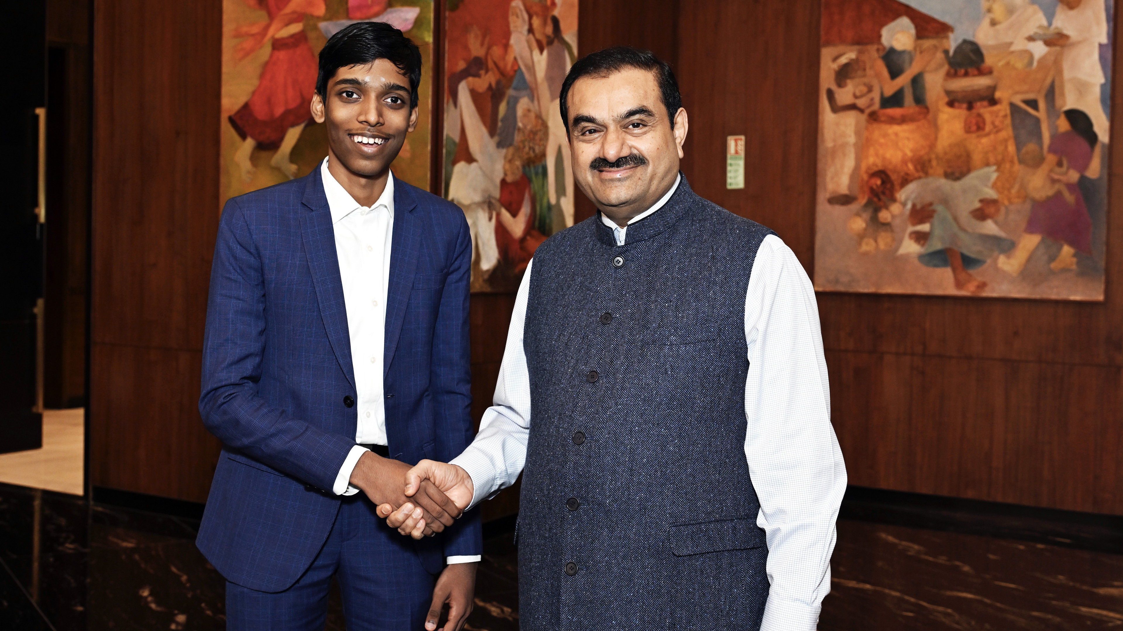 Gautam Adani and a young man in business suits shaking hands and smiling for a photograph.