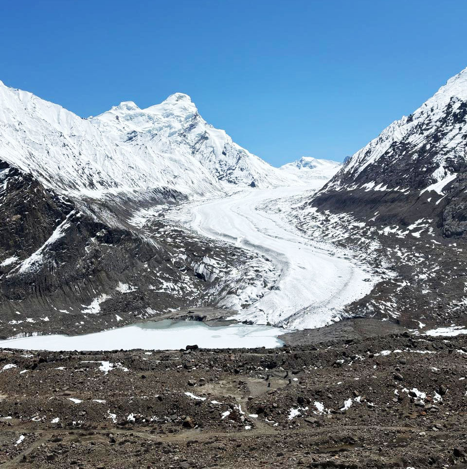 A vast winding glacier descends through a rugged valley, flanked by snow-capped mountains, meeting a frozen lake.