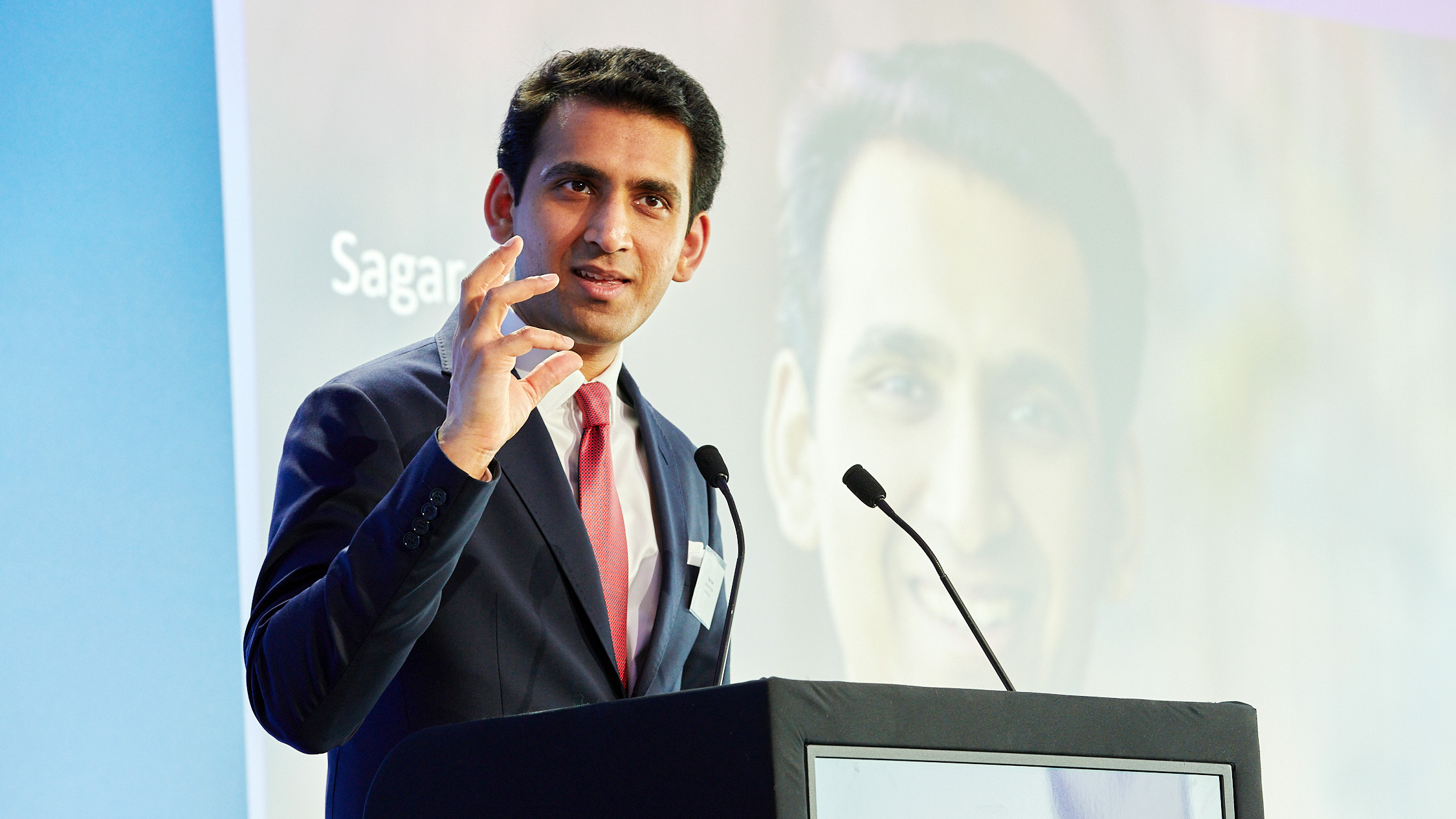 A man in a suit and red tie speaks at a podium, gesturing with his hand during a presentation.