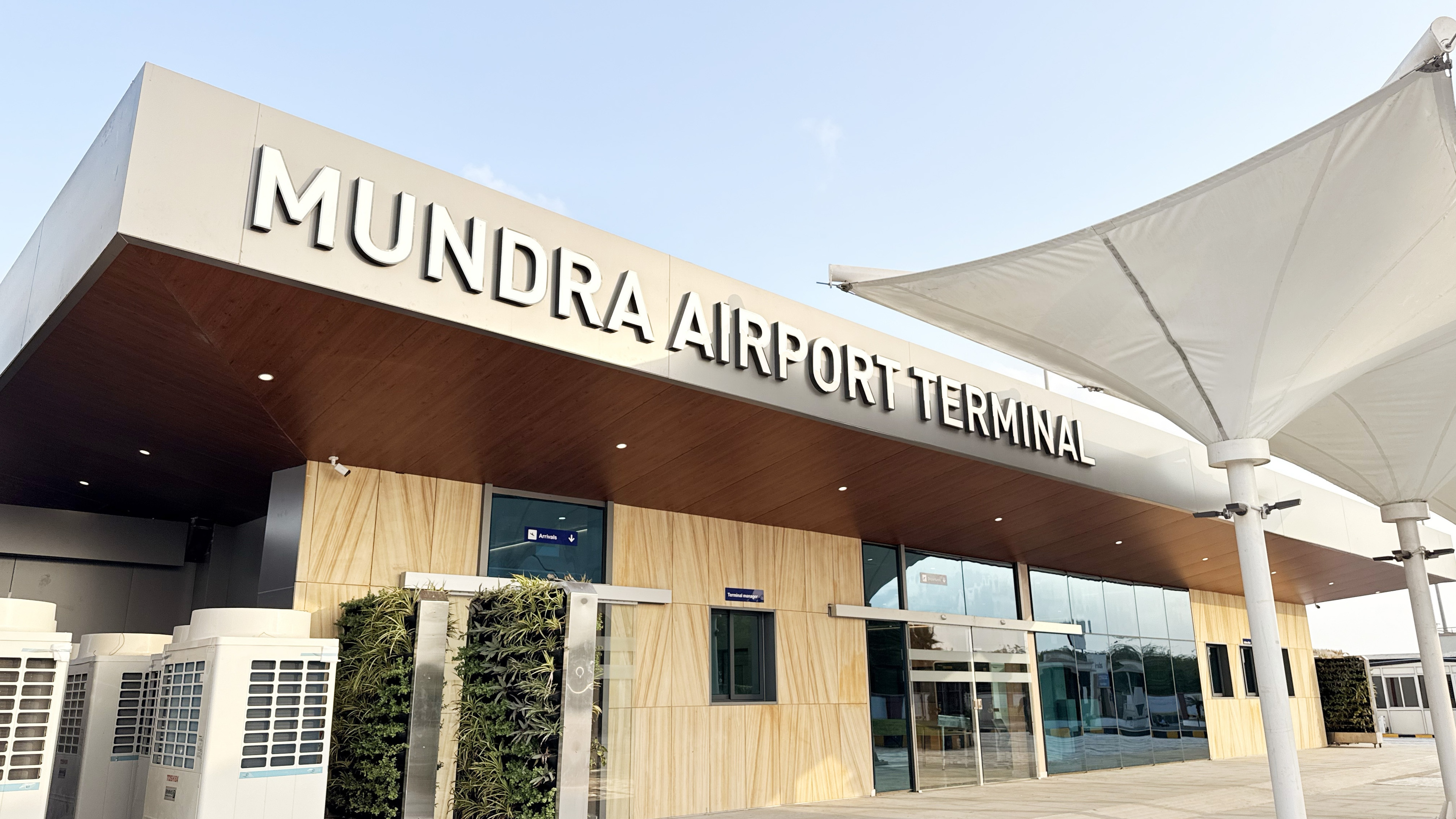 Exterior view of Mundra Airport Terminal building with its prominent sign, modern architecture, and outdoor canopies.