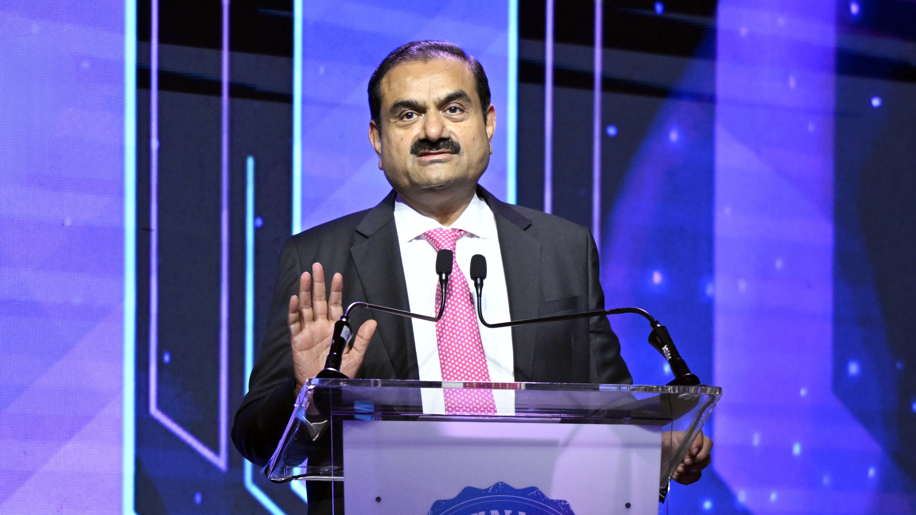 Industrialist Gautam Adani speaks at a podium, raising his hand, against a blue background.