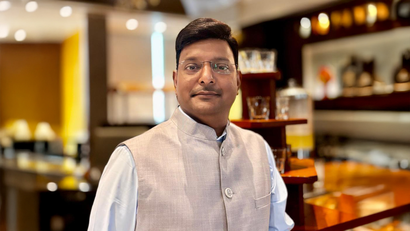 A man wearing a light blue shirt and a light grey Nehru jacket, standing indoors. He is looking at the camera.