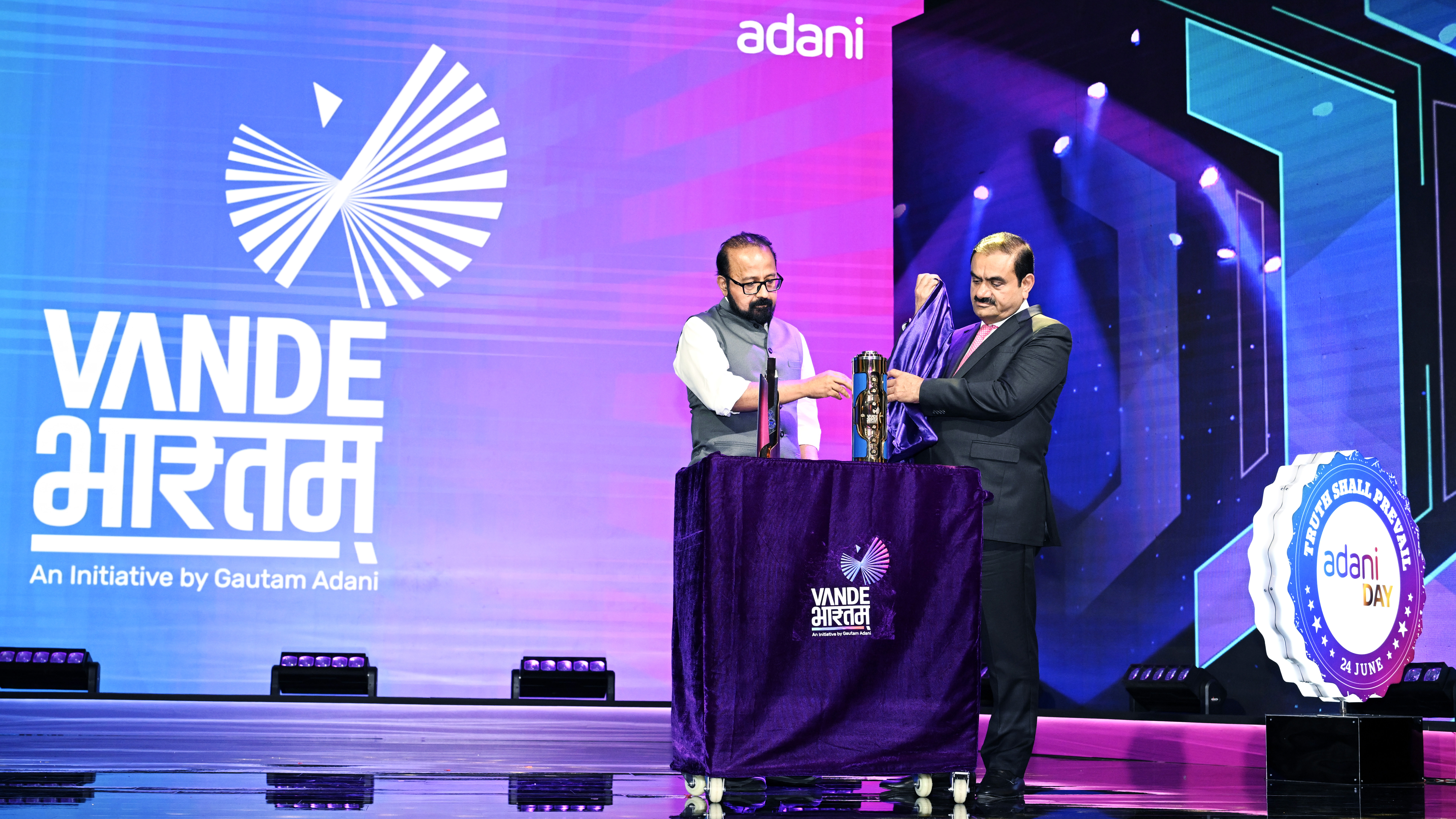 Two men on stage unveiling an object. A large screen behind them shows "Vande Bharat" and "Adani" logos.
