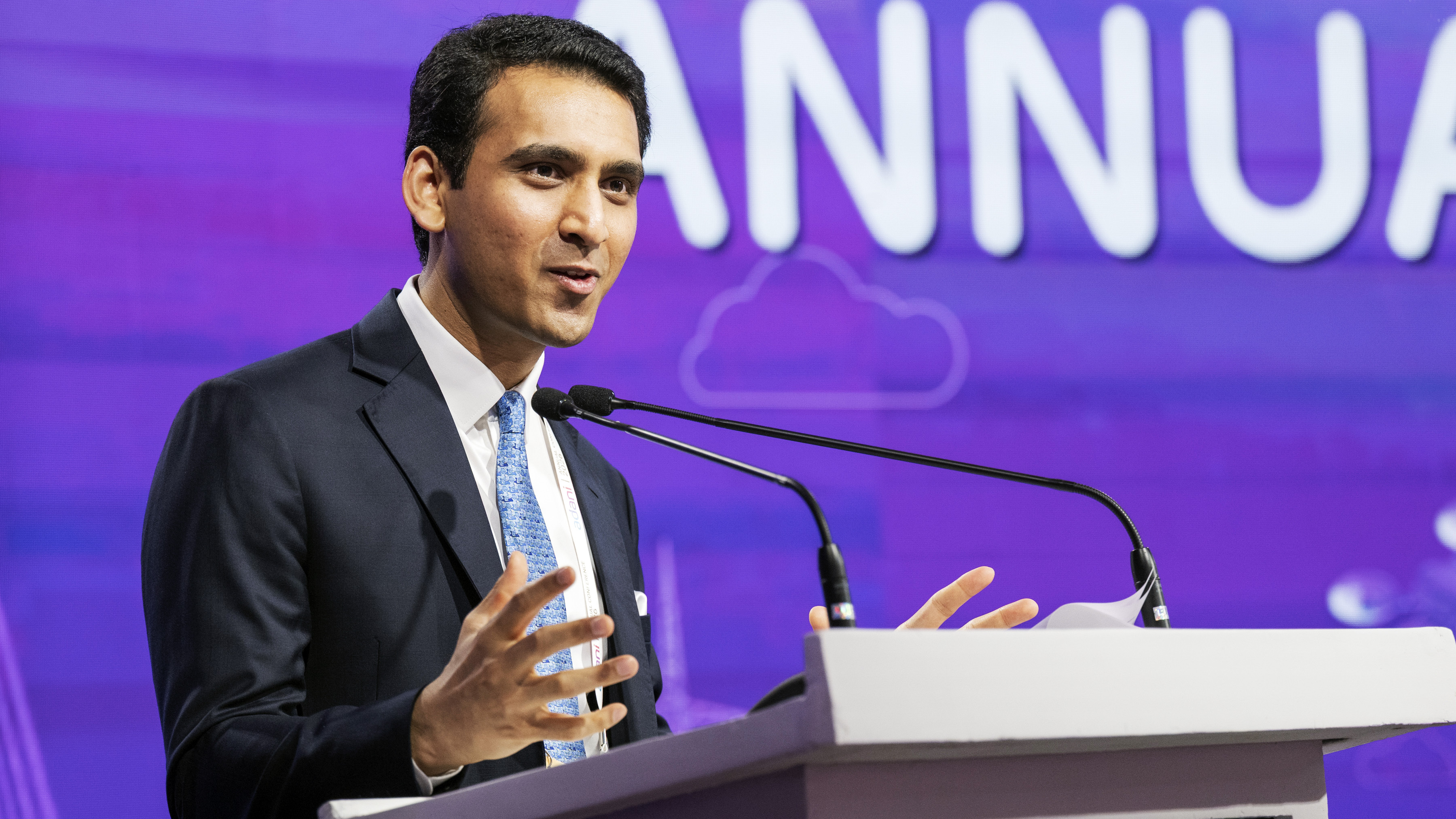 A man in a suit and tie speaks at a podium with microphones, gesturing with his hands.