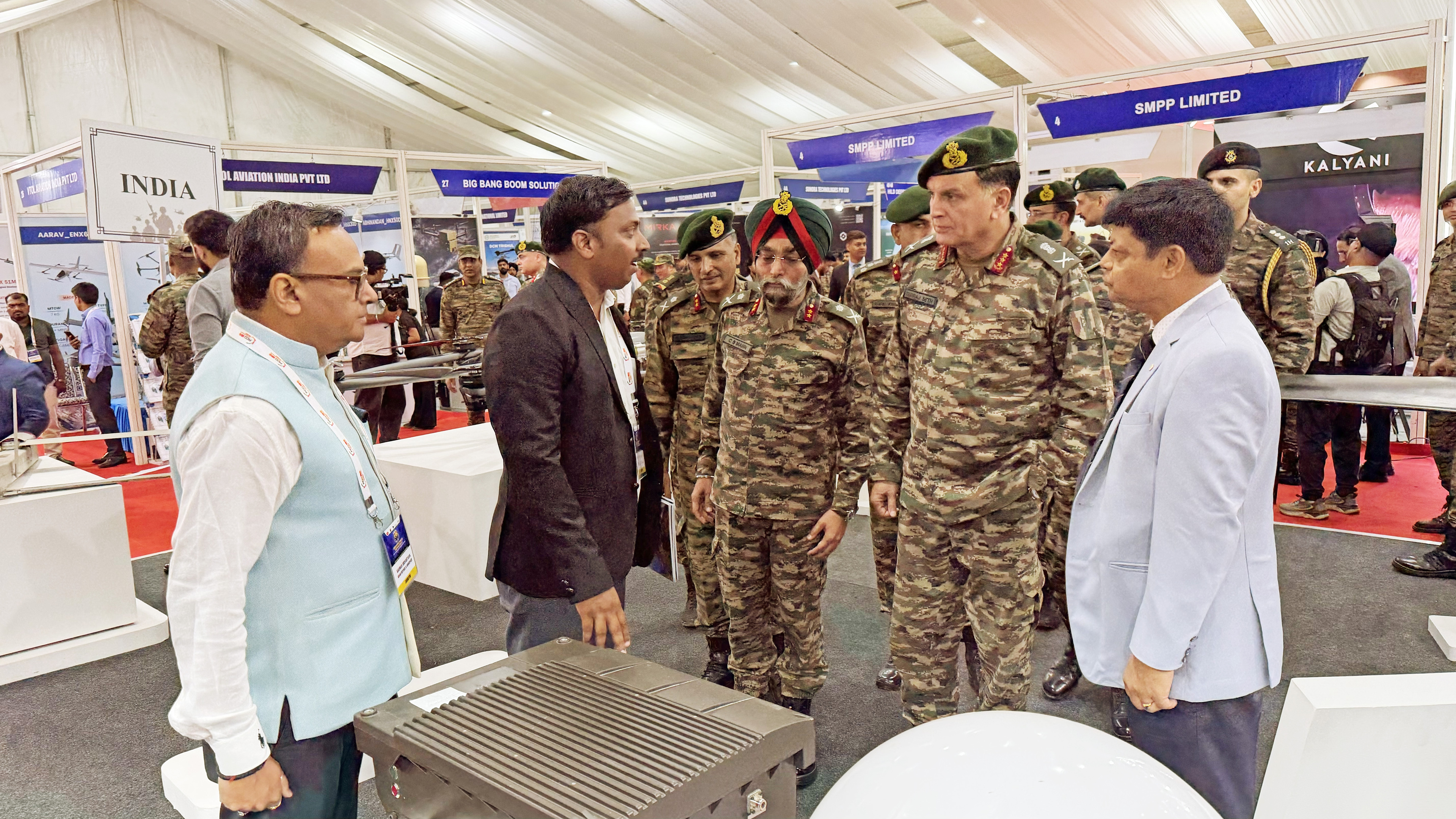 A group of military officers and civilians discuss a device at a defense exhibition booth in India.