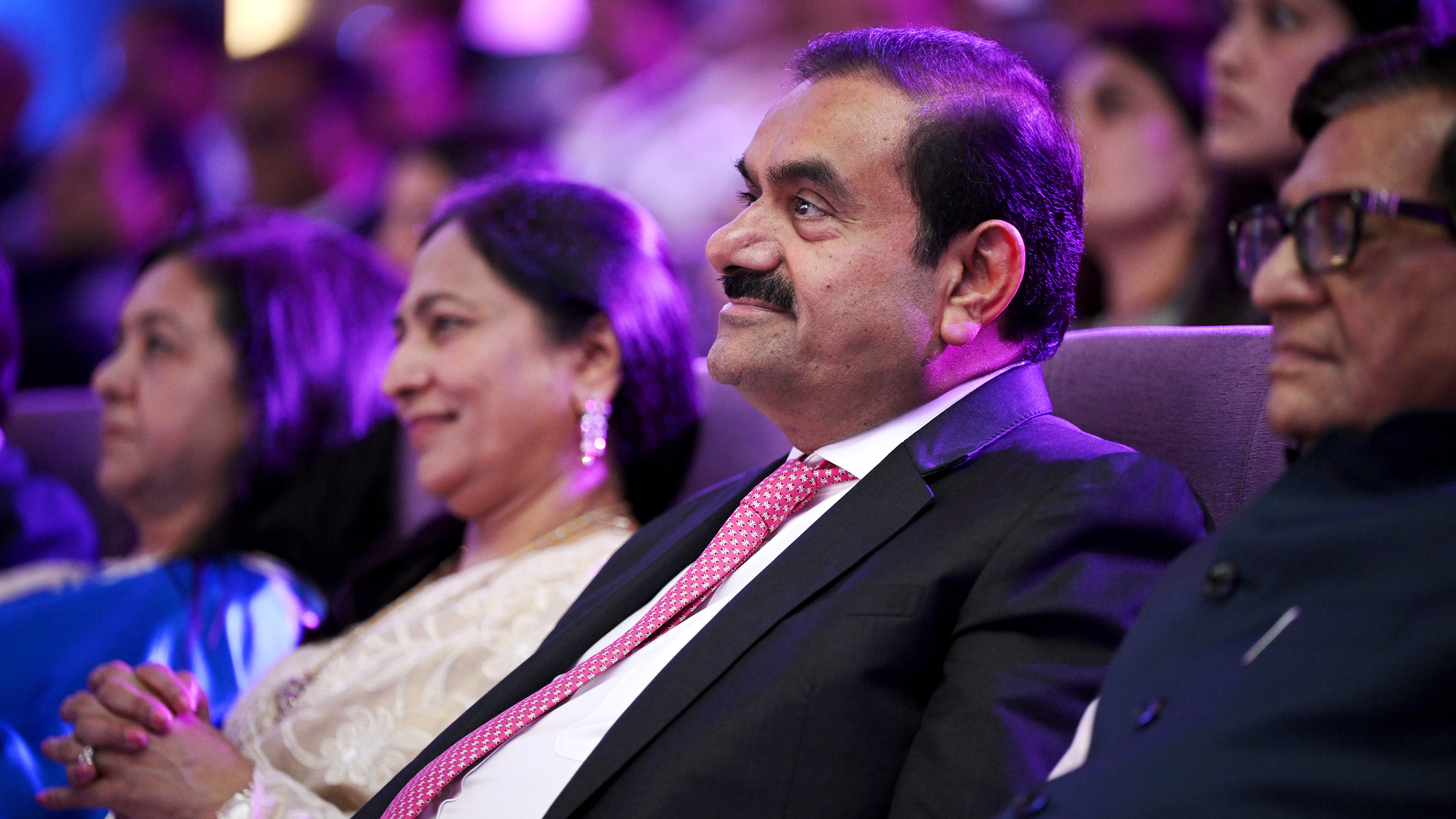 Gautam Adani seated at an event with other attendees, under vibrant purple lighting.