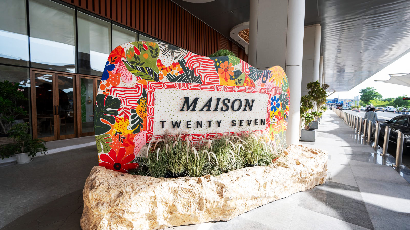 A vibrant, colorful sign for Maison Twenty Seven with a rock planter and grass, at a modern building entrance.
