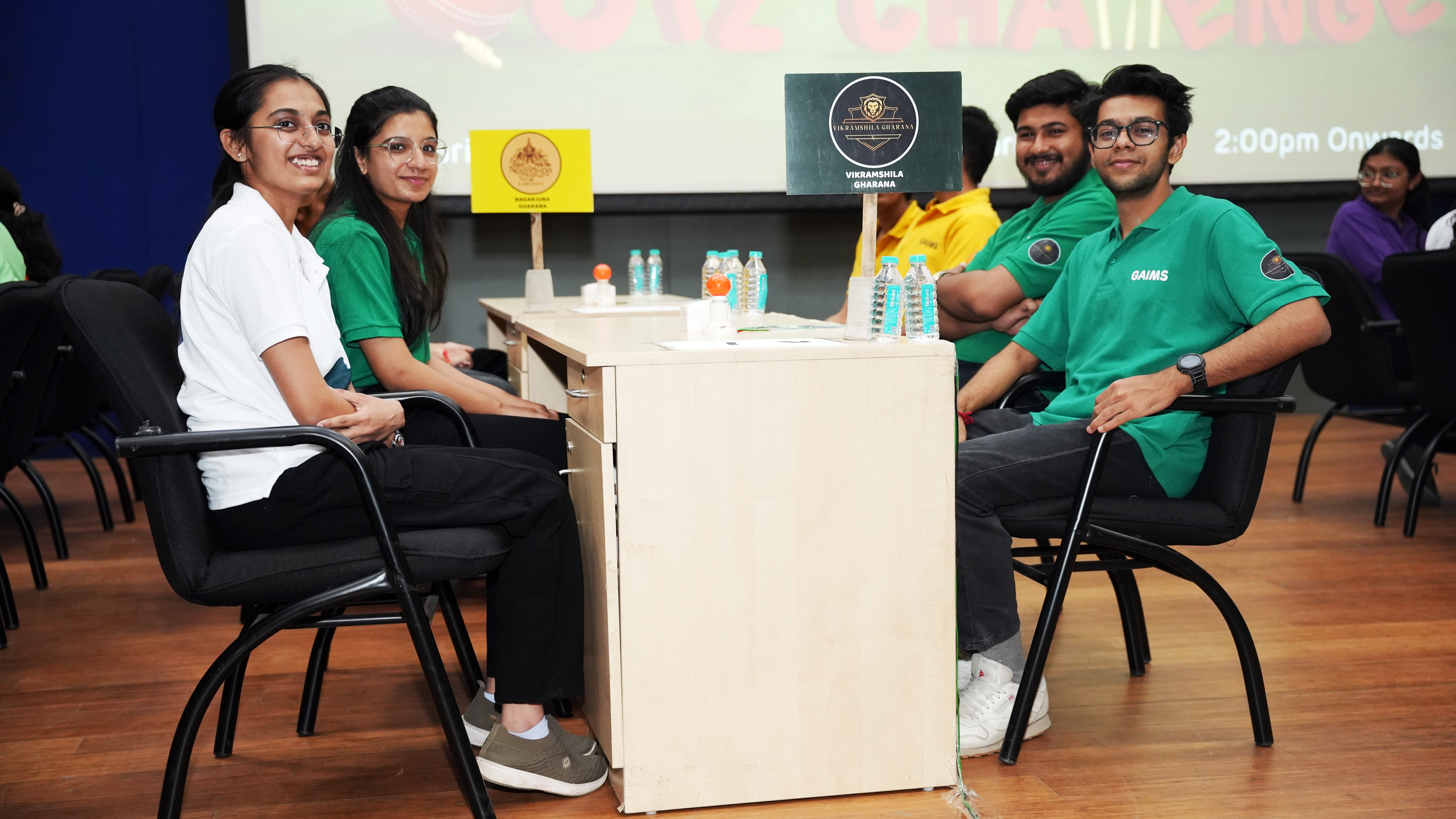 Four students in green and white shirts sit at a table during a quiz or challenge event, smiling at the camera.
