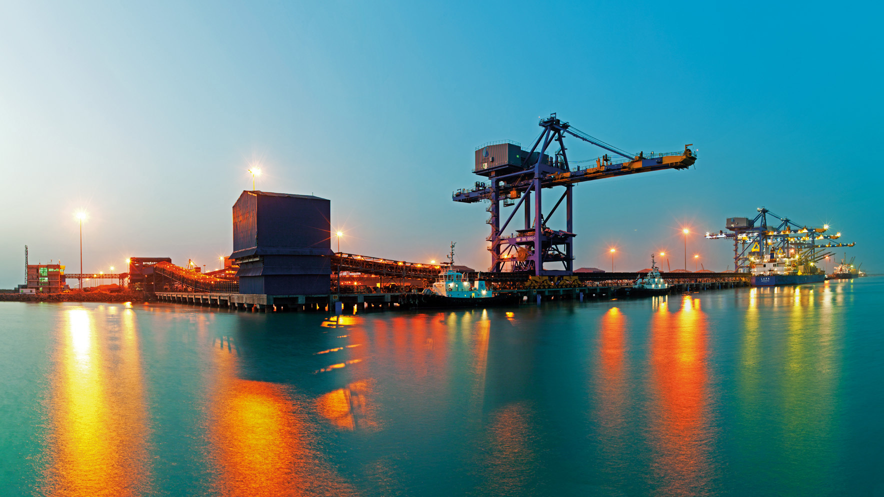 Night view of a bustling industrial port with brightly lit structures, cranes, and a ship reflecting on calm water.