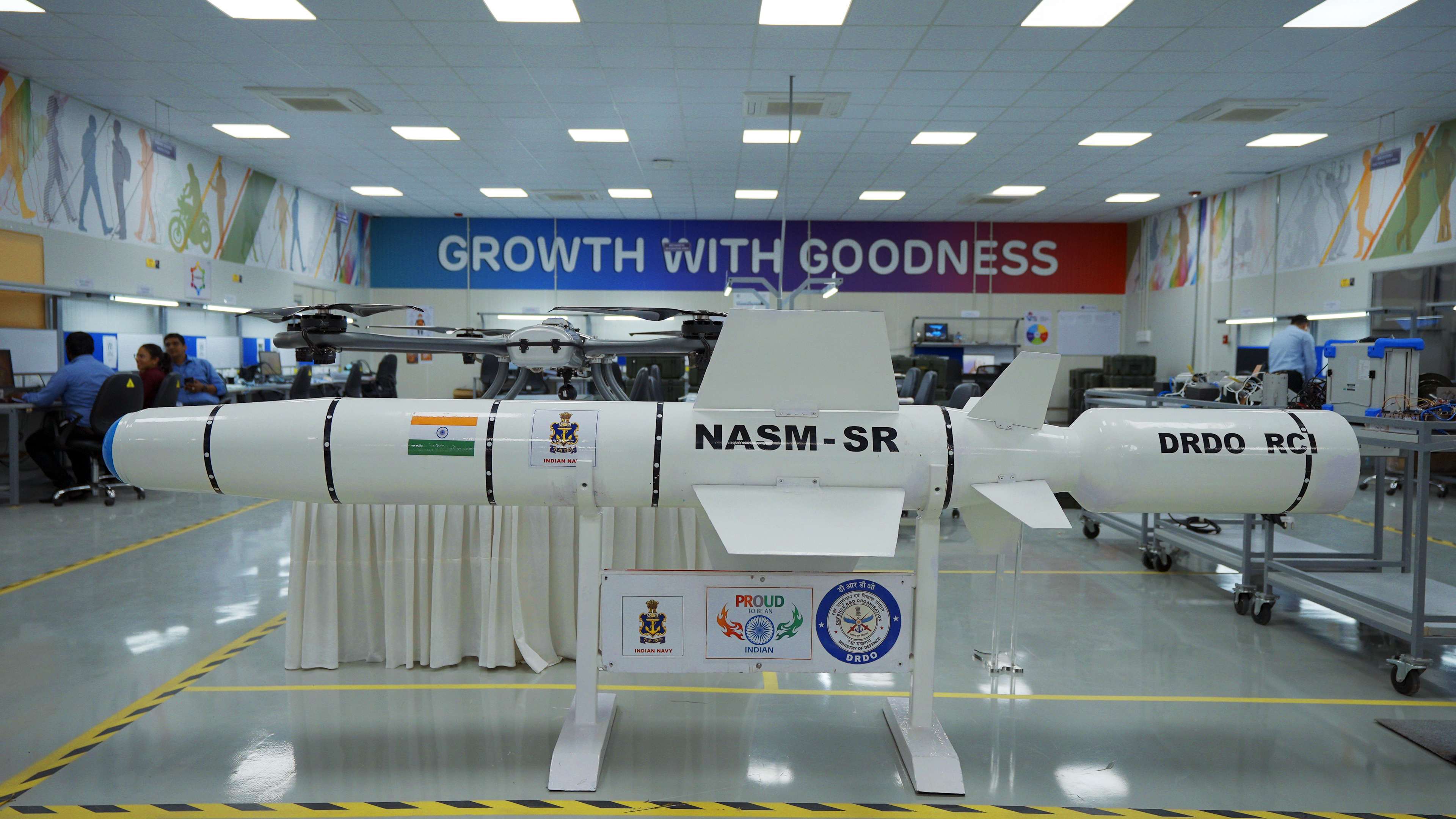 A white NASM-SR missile model with an Indian flag and a drone atop it, displayed in a laboratory.