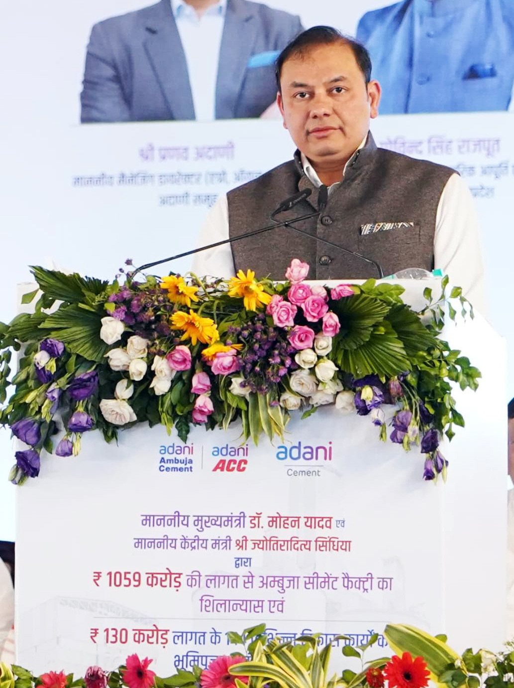 Man speaks at Adani Cement factory groundbreaking event, podium with flowers.