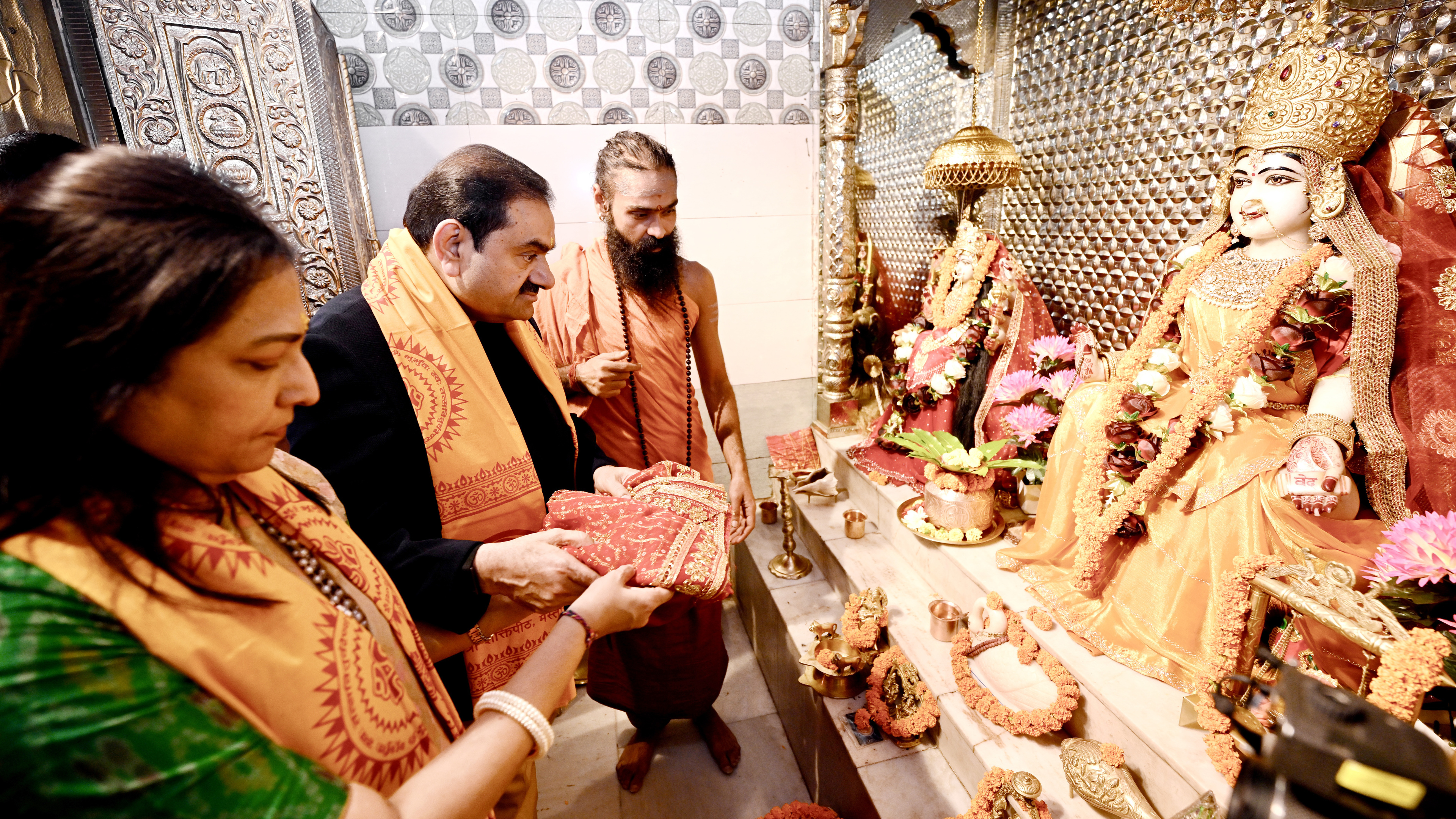 Gautam and Priti Adani performing puja with a priest in a temple, presenting offerings to deities.