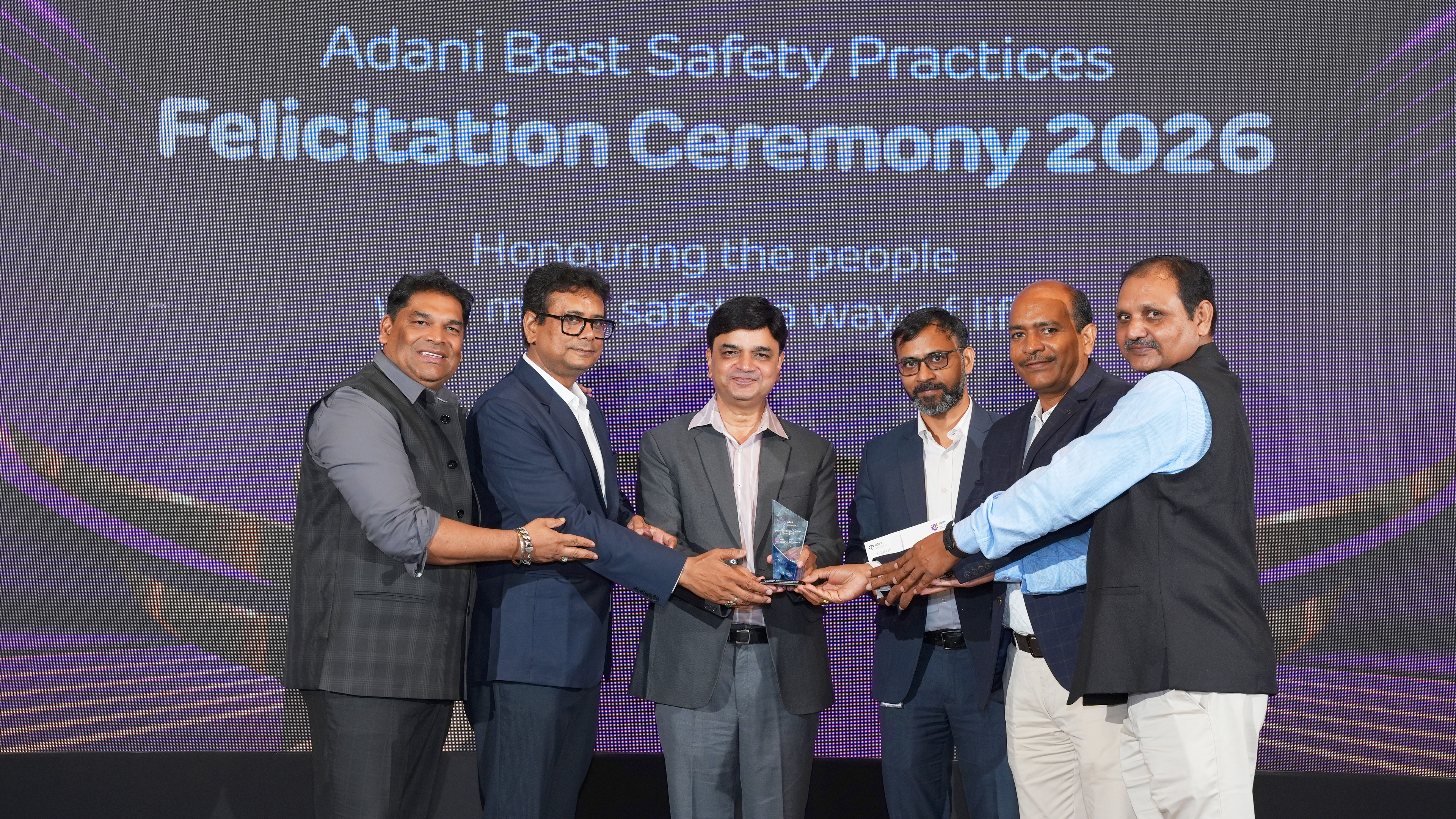 Six men on stage accepting an award at the Adani Best Safety Practices Felicitation Ceremony 2026.