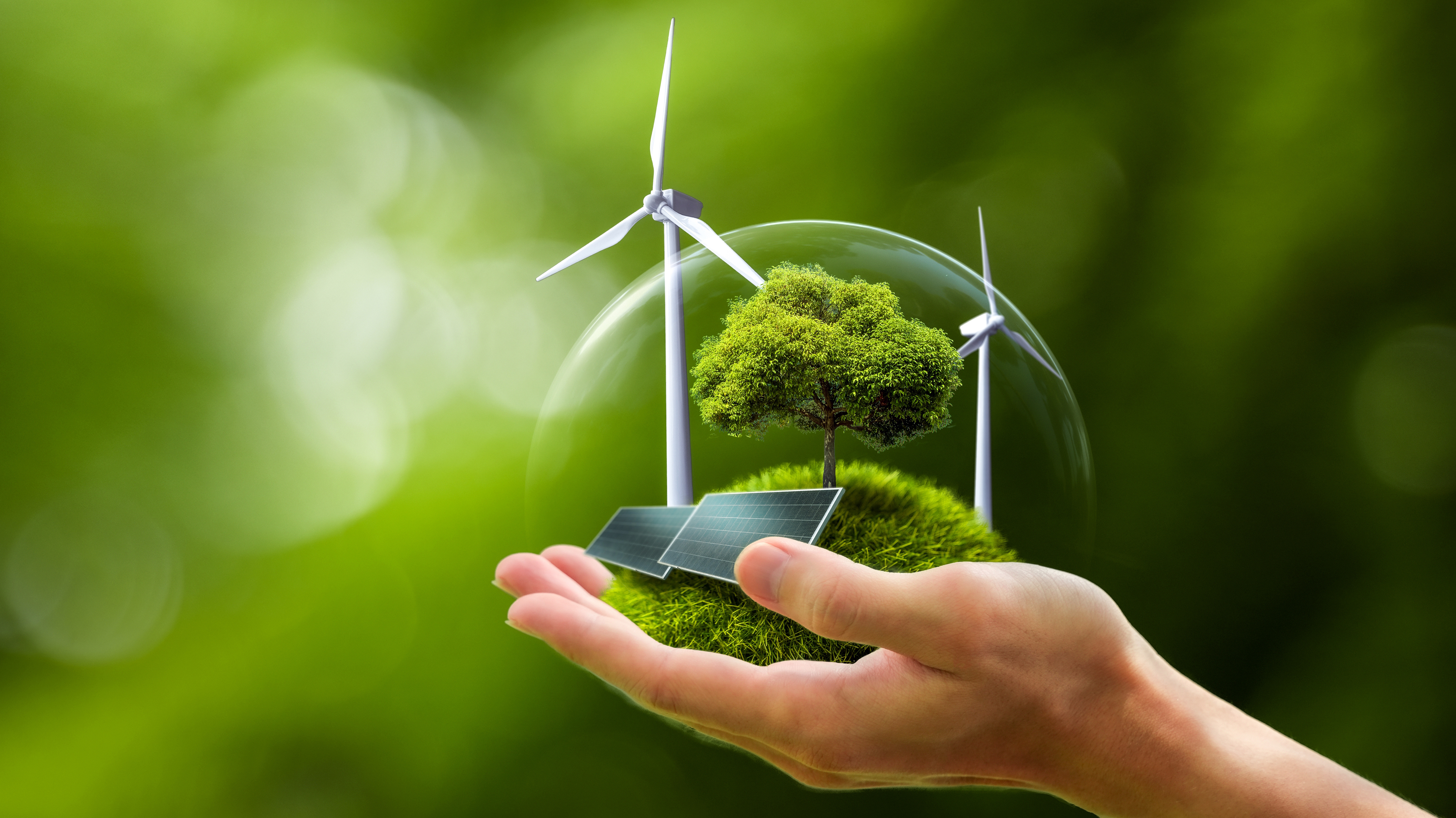 Hand holds a bubble with wind turbines, solar panels, and a tree on grass, symbolizing clean energy.