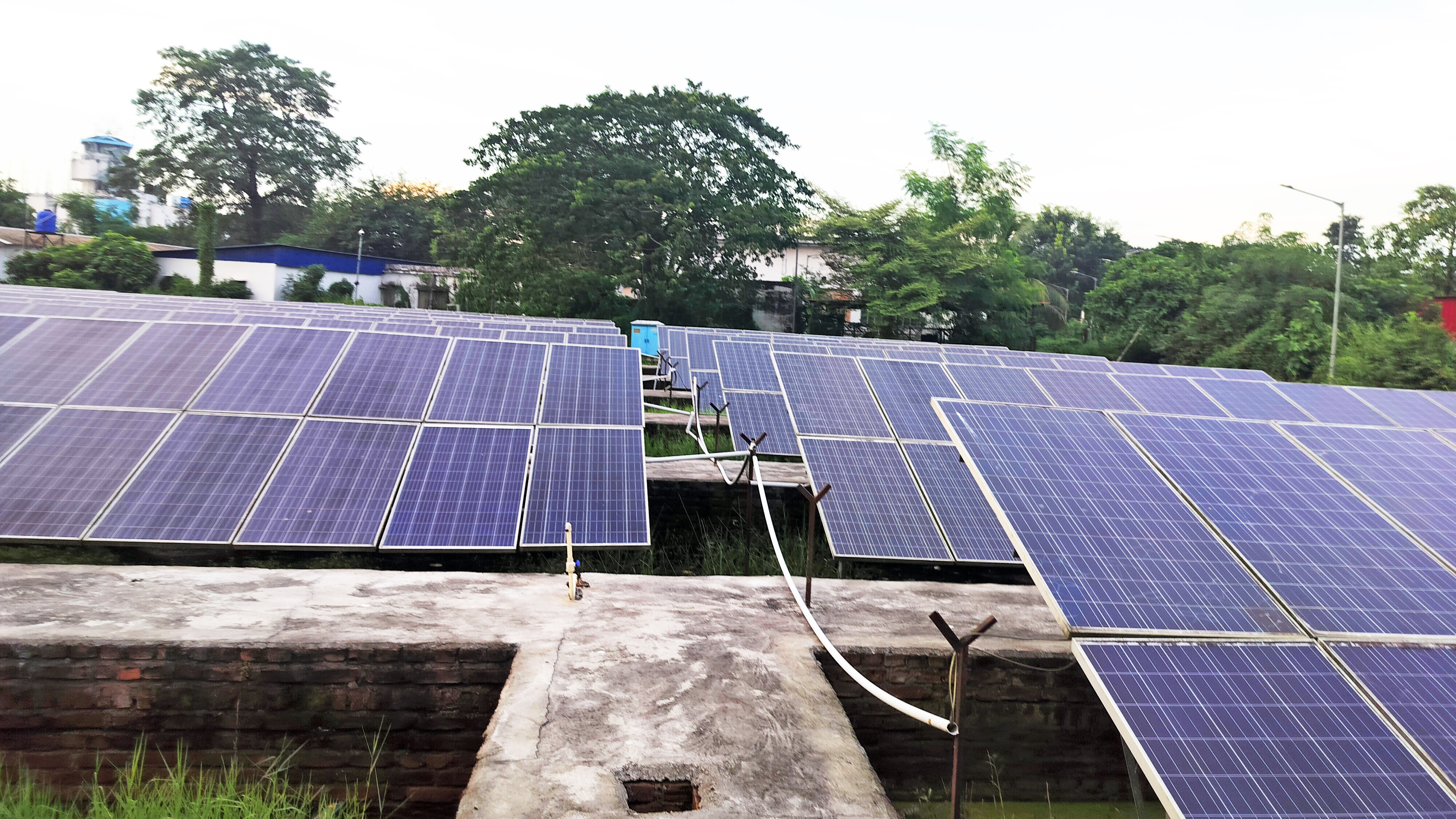 Expansive field of solar panels on a concrete base with trees and buildings in the background under a bright sky.