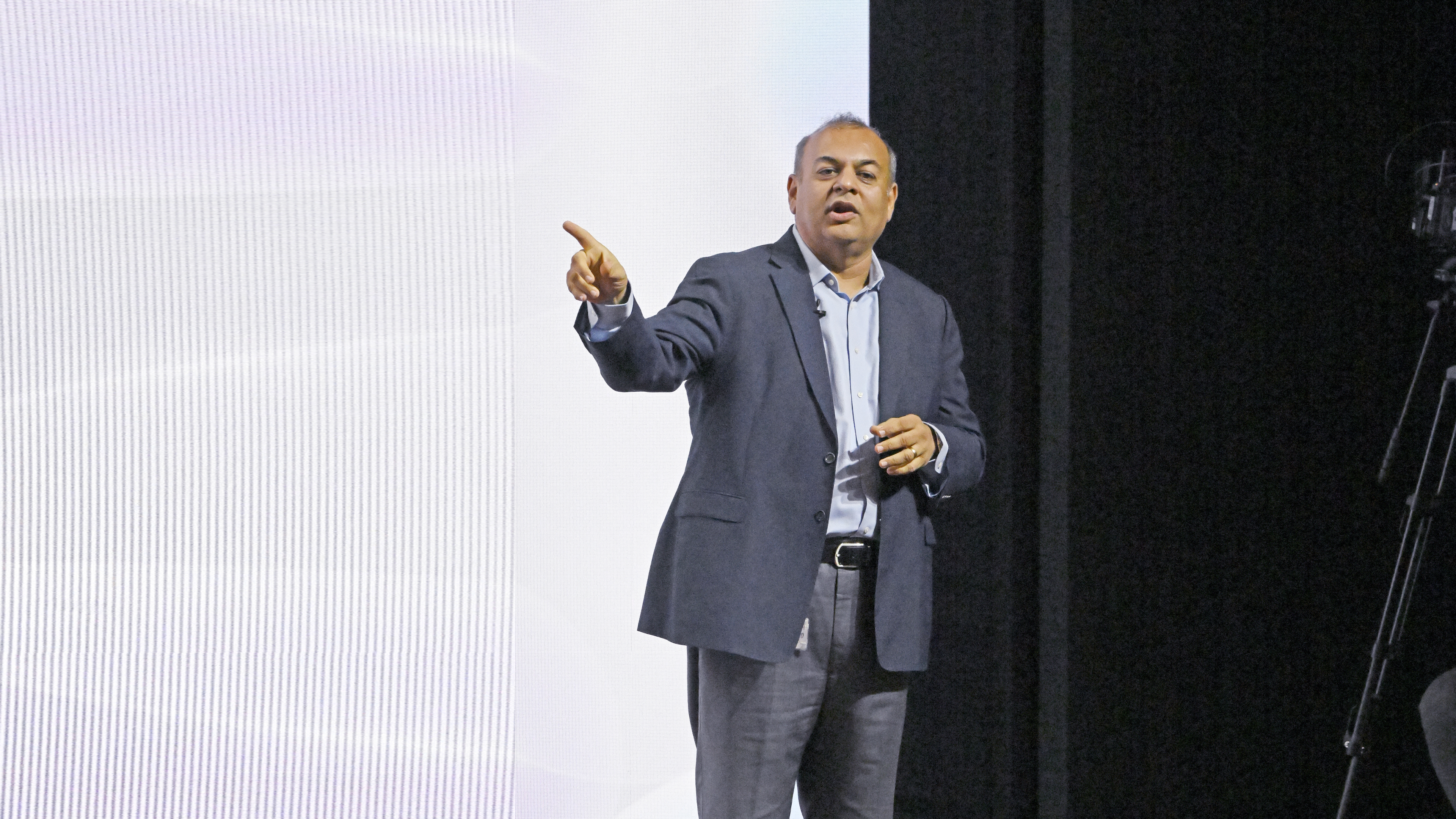 A man in a suit presenting on stage, pointing to his left with a confident expression.