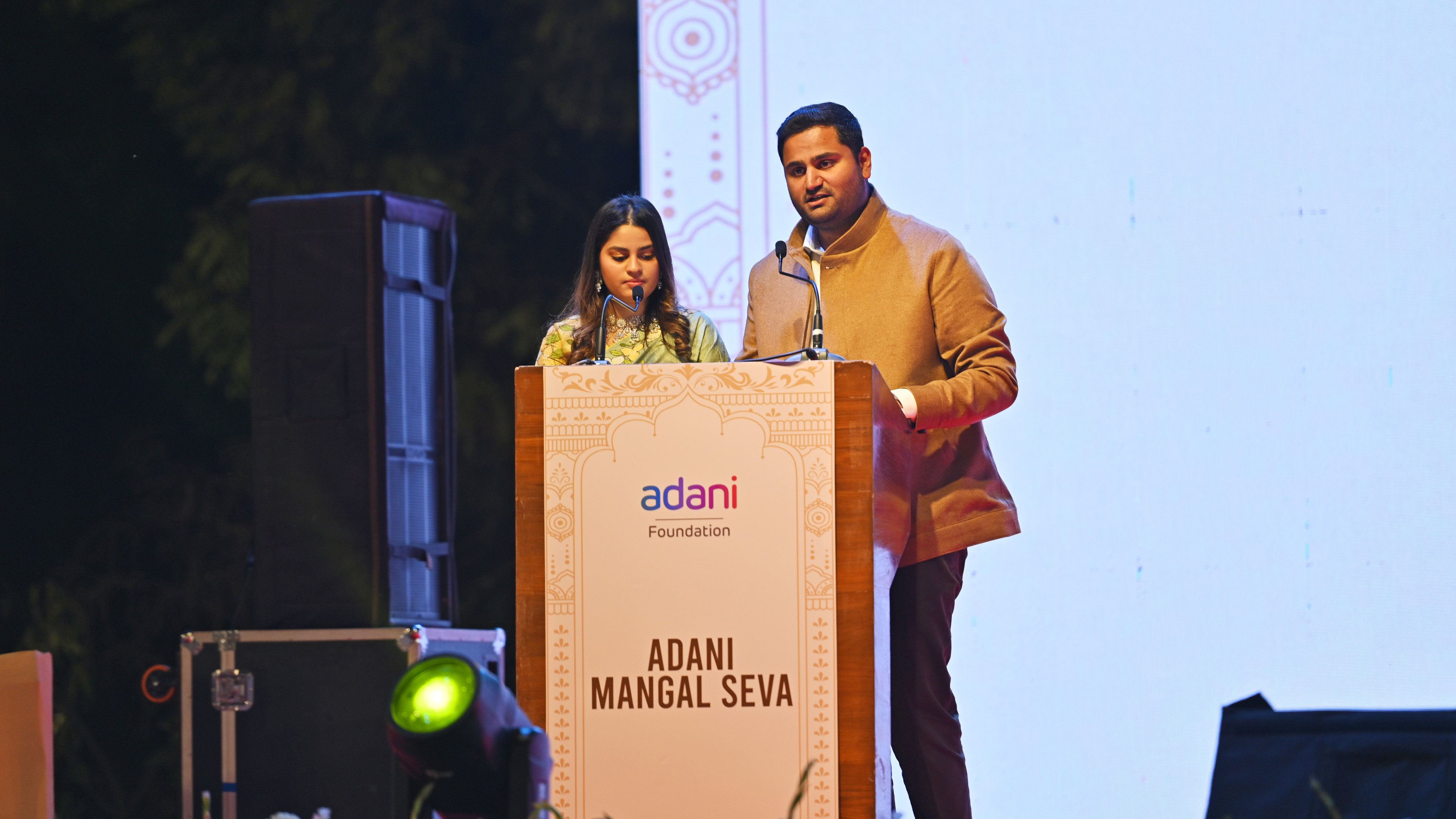 A man speaks from a podium with the Adani Foundation logo, while a woman stands quietly behind him.