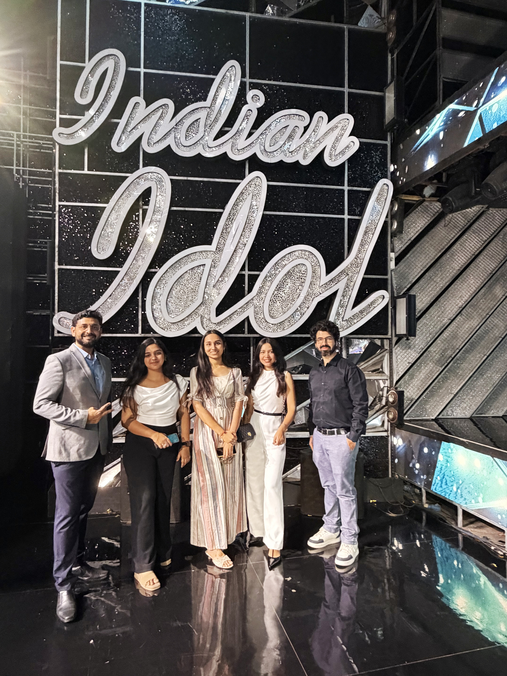 Five people, two men and three women, smile in front of the glittering 'Indian Idol' logo on a stage.