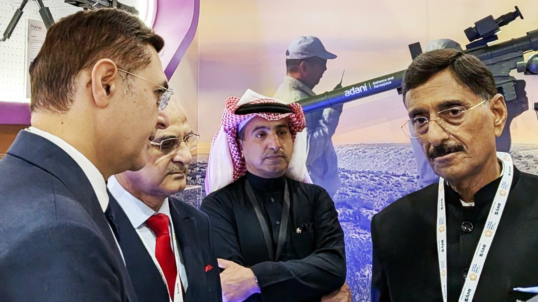 Four men, including one in a keffiyeh, are engaged in discussion at a defense exhibition booth.