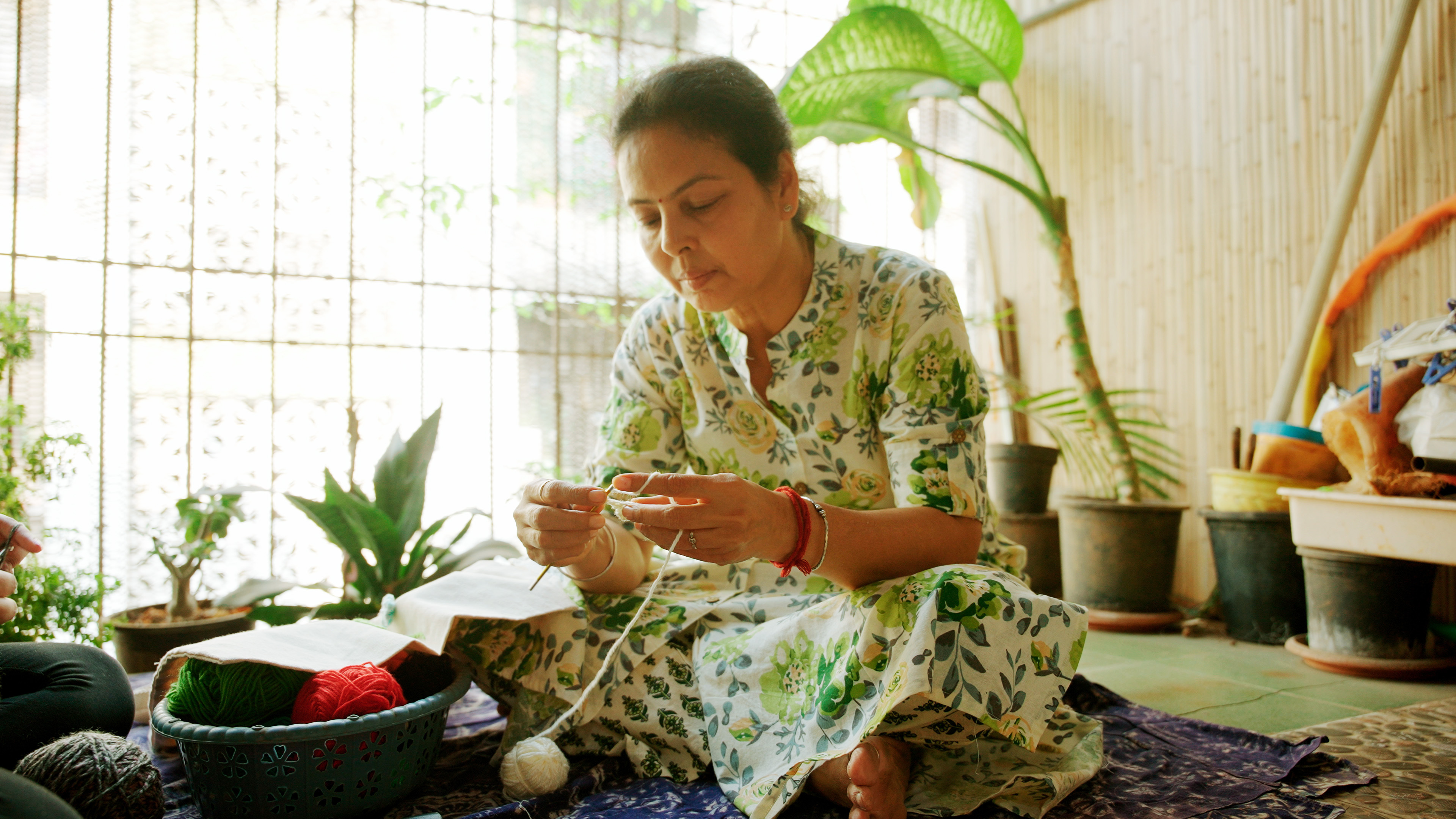A woman sits on the floor, focused on crocheting with white yarn, a basket of colored yarn nearby.