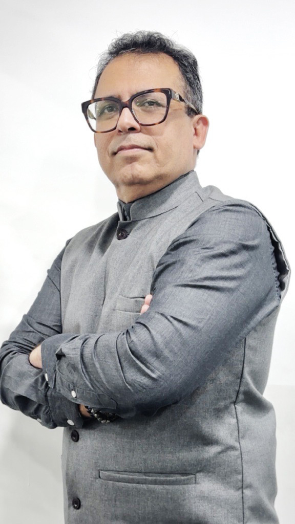 A man in a grey shirt and vest, wearing glasses, stands with arms crossed, looking confidently at the camera.