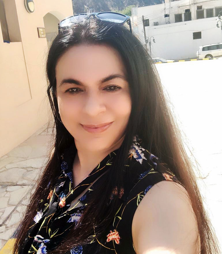 Smiling woman with long dark hair in a floral top, outdoors on a sunny day with buildings and mountains.