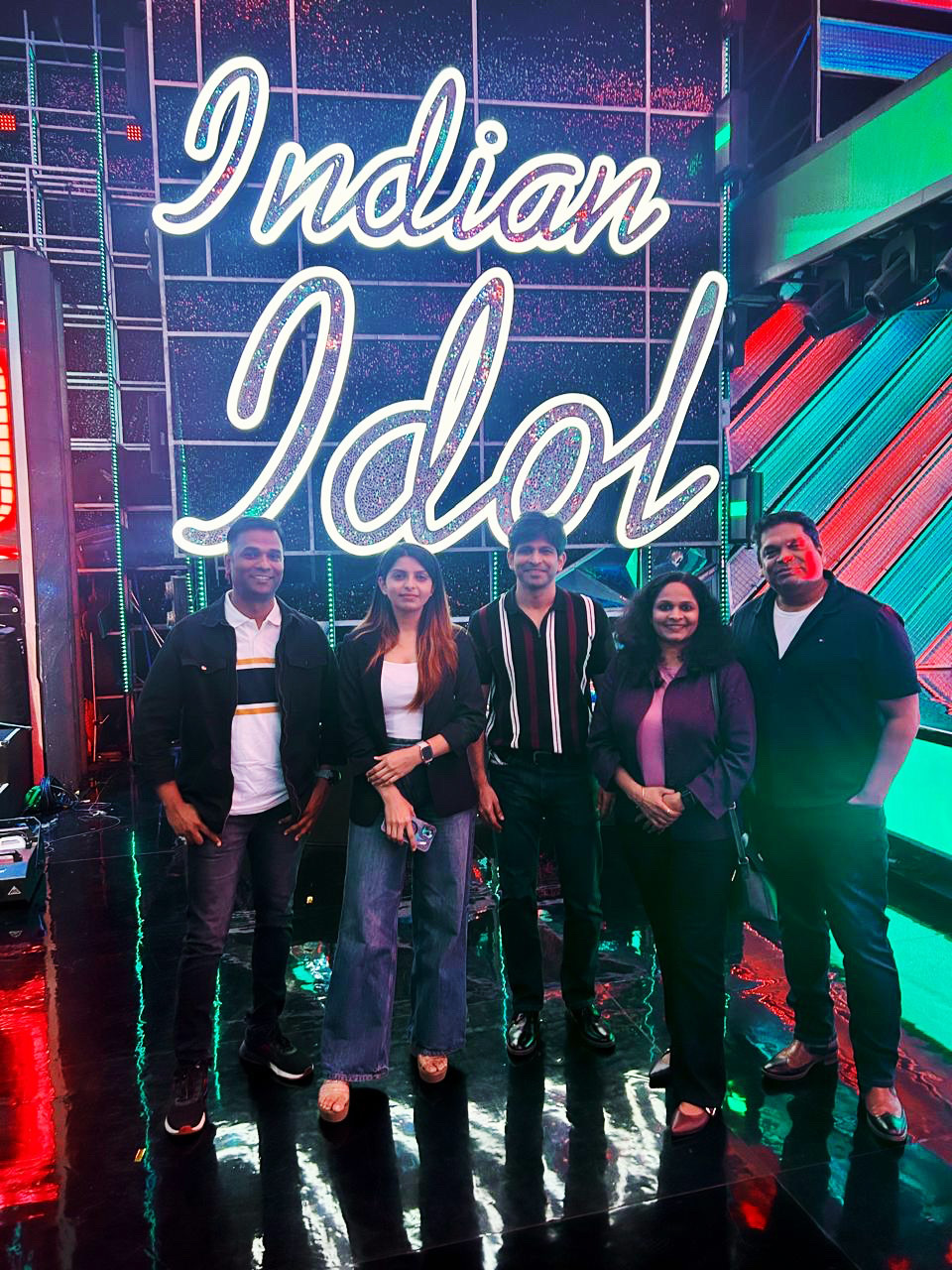 Five people pose in front of a glowing "Indian Idol" sign on a stage, lit by colorful lights.