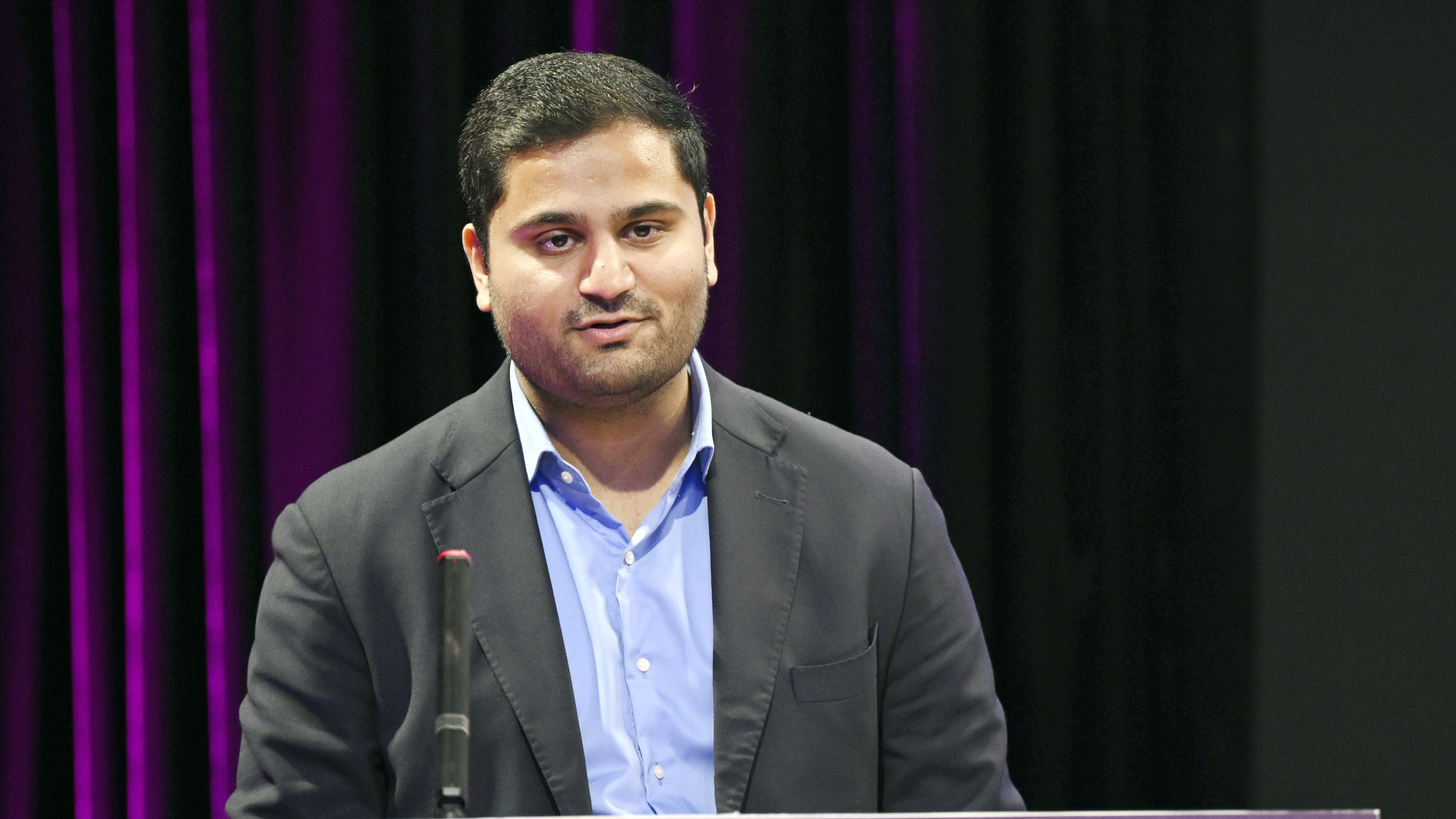 A man with dark hair and a beard, wearing a blue shirt and suit jacket, speaks at a podium.
