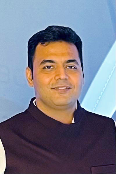 A man with dark hair wearing a dark vest over a white shirt smiles at the camera against a light blue background.