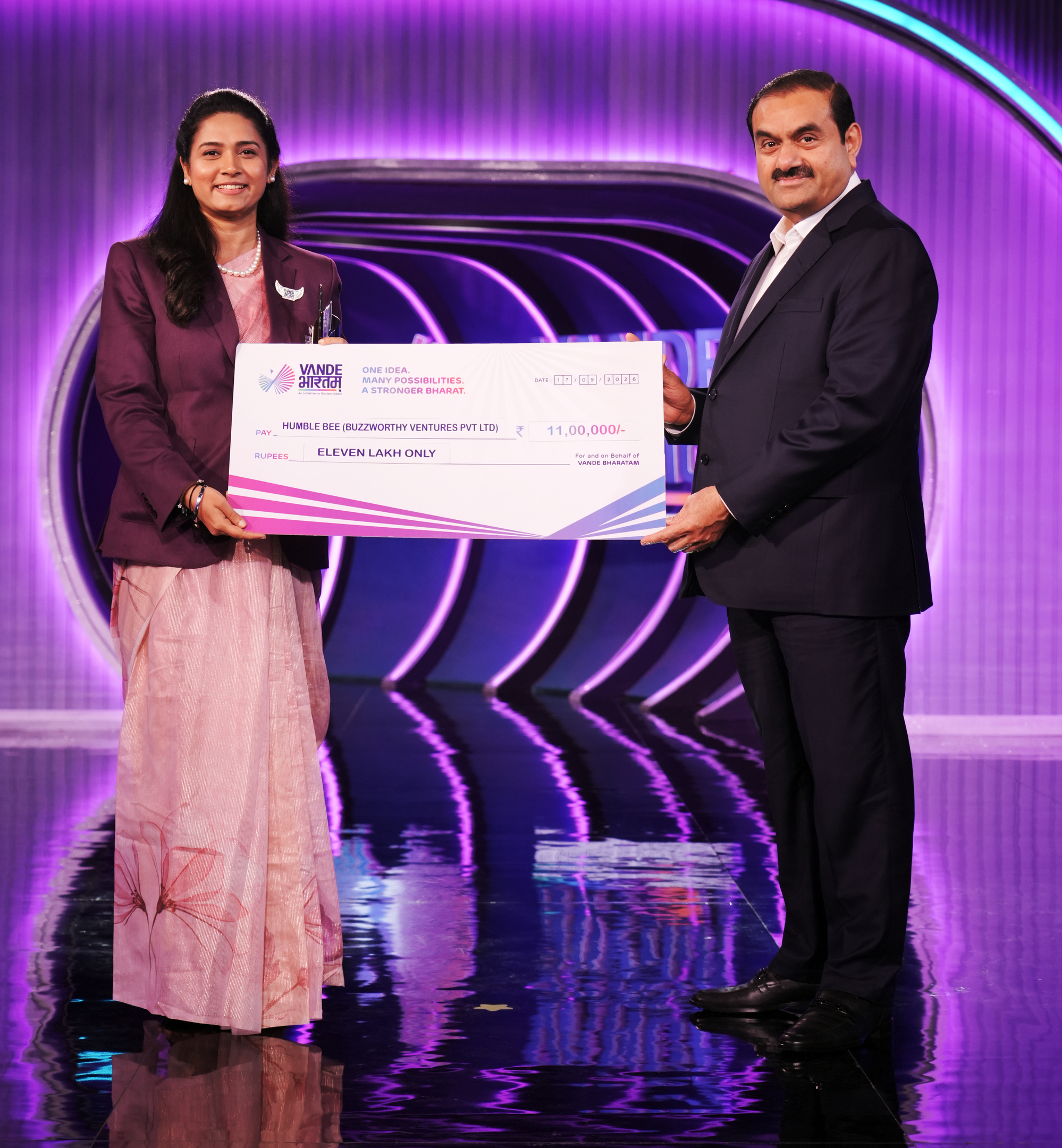 Two people, a woman and a man, holding a large check for 11,00,000 INR from Vande Bharatam.