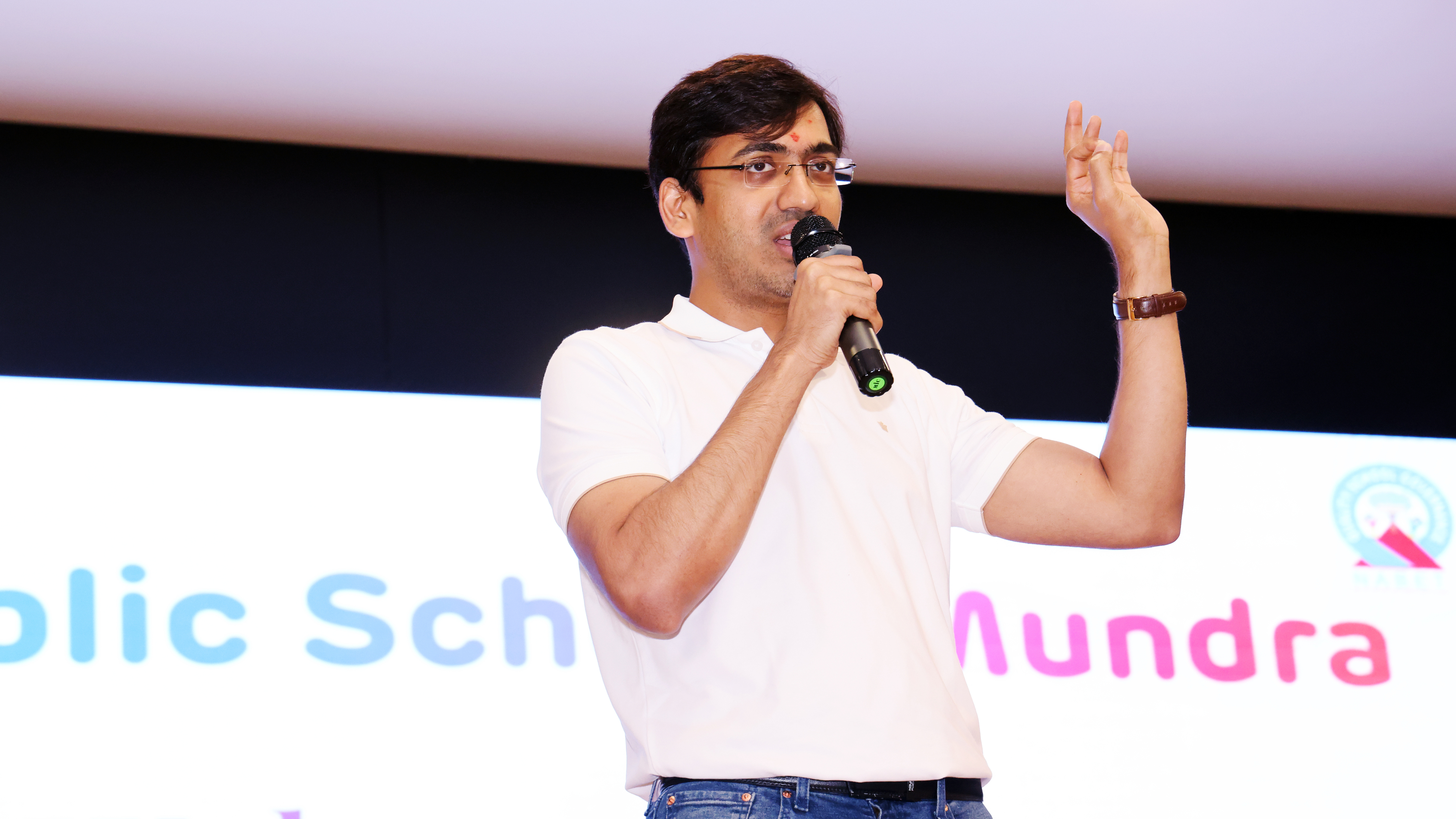A man in a white polo shirt and glasses speaks into a microphone, gesturing with his right hand.