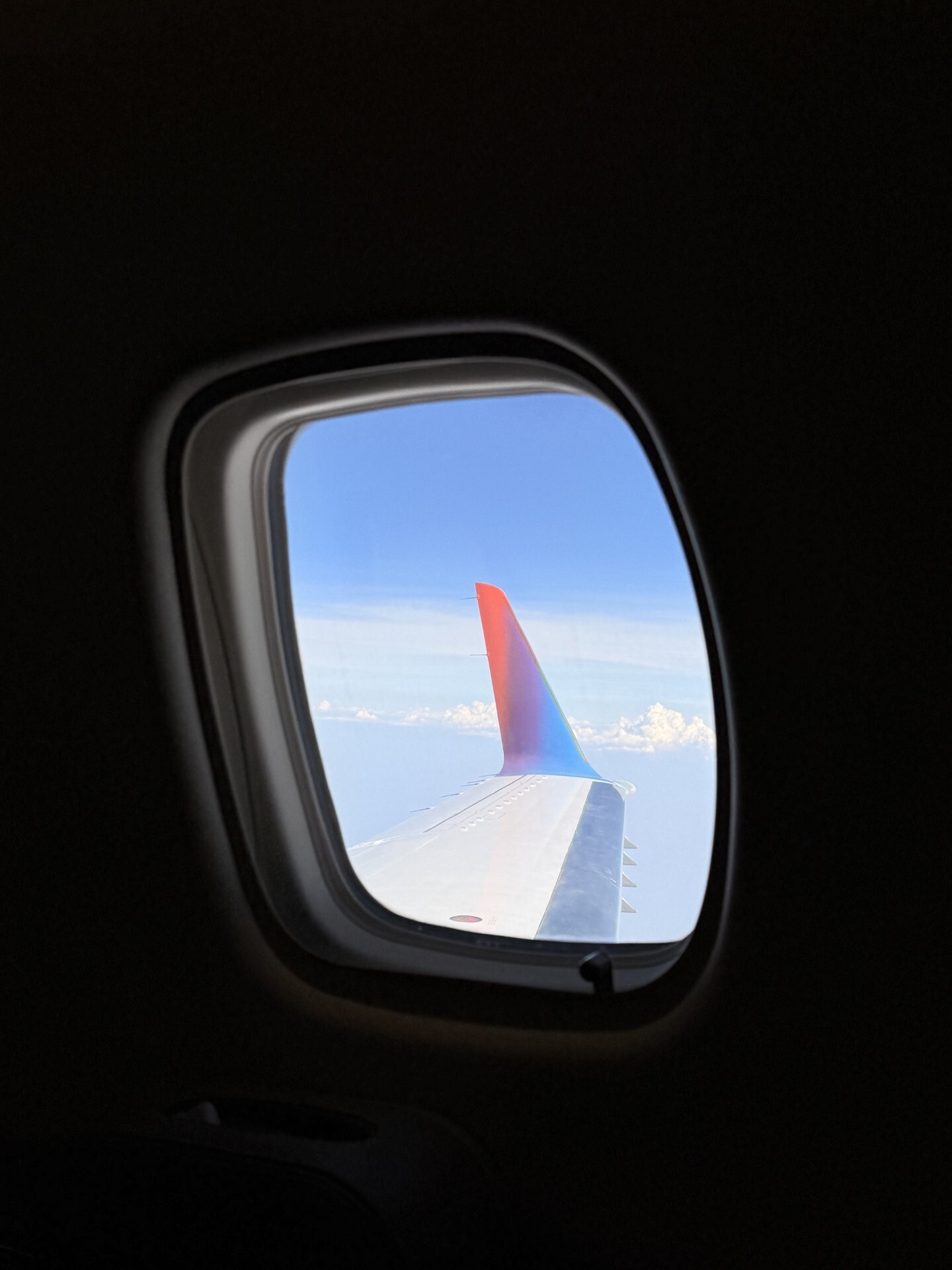 View from an airplane window showcasing the wing with a colorful tip (red, orange, blue) against a blue sky with clouds.