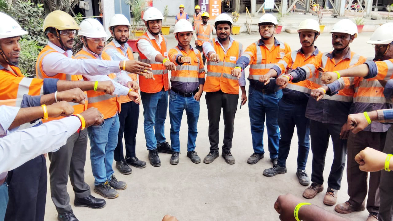 A group of construction workers in hard hats and orange vests stand in a circle with arms extended and fists forward.