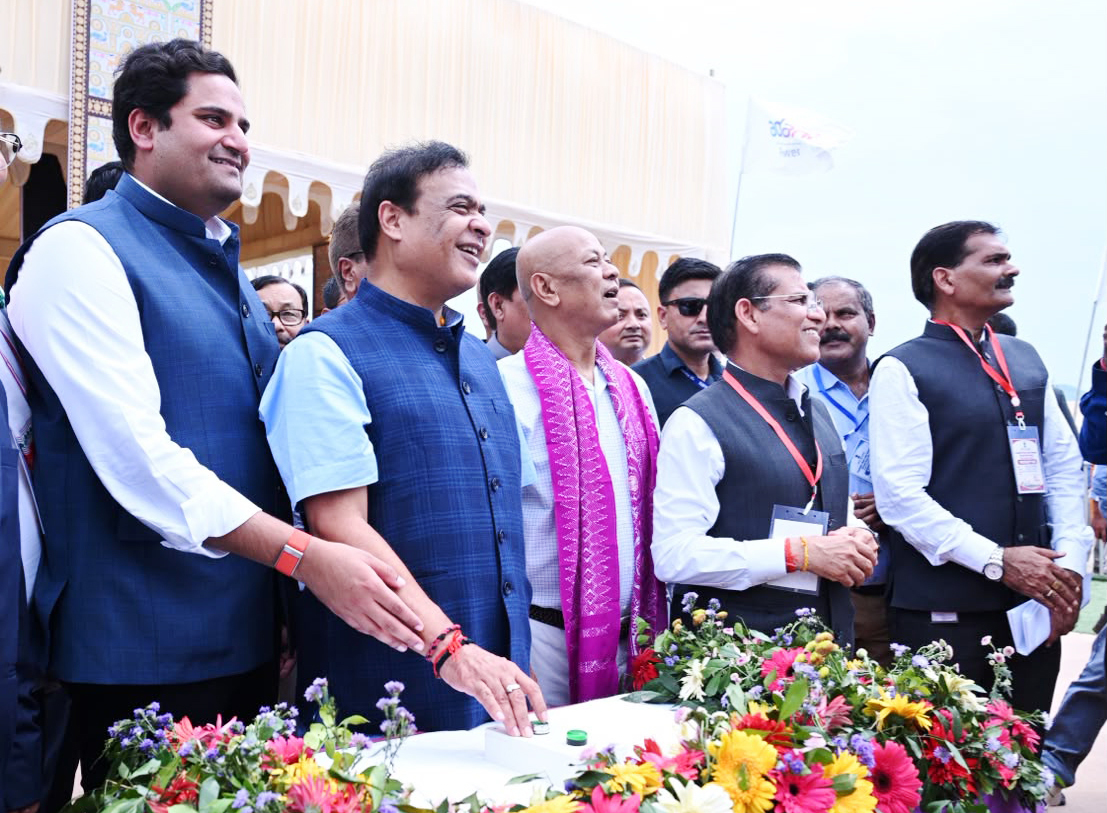 Group of smiling men, including politicians, at an outdoor event, one pressing a button on a flower-decorated table.