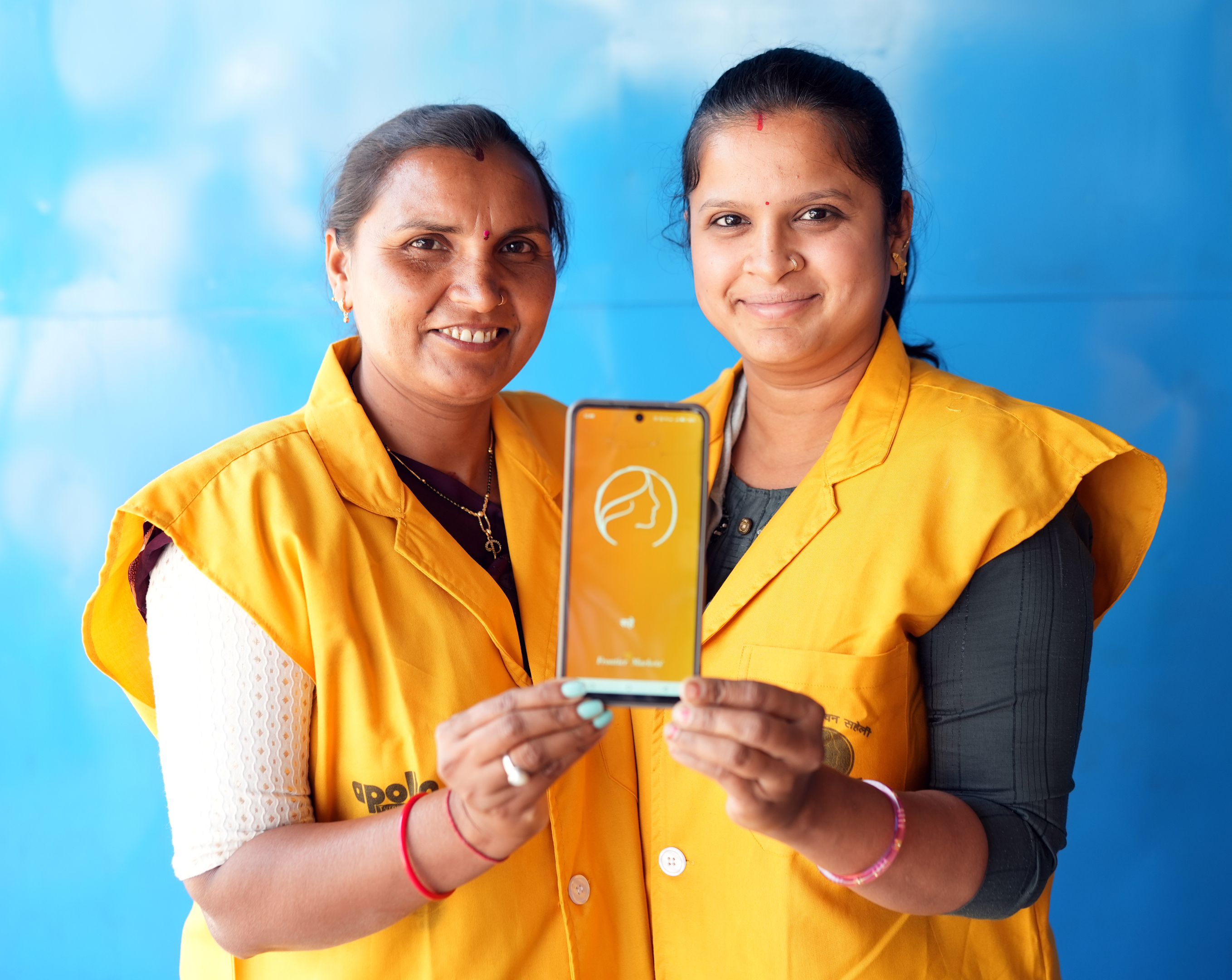 Two smiling women in yellow vests hold up a smartphone displaying an app logo, against a blue background.