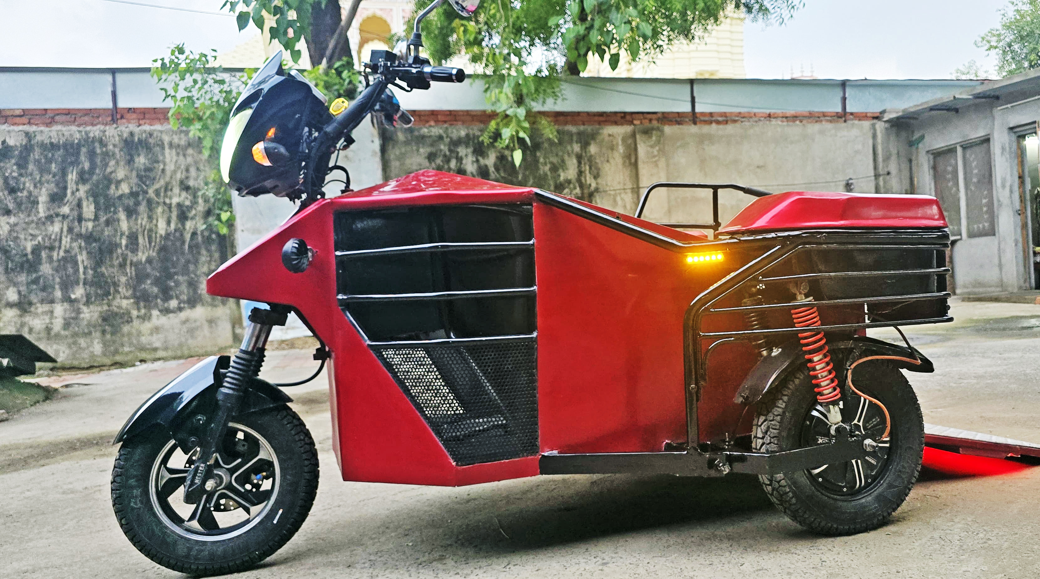 A red electric delivery tricycle with a cargo bed on a paved surface, against a wall and trees.
