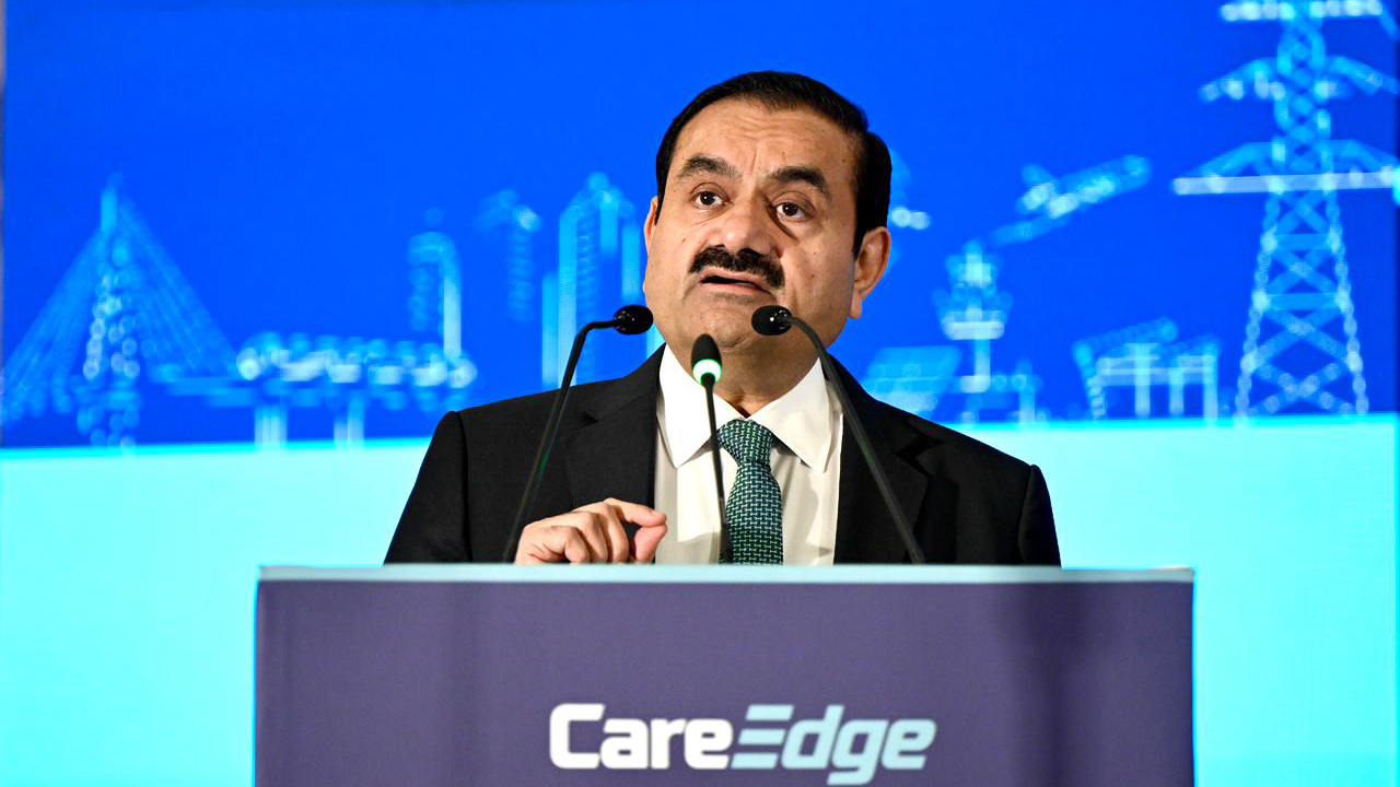 Gautam Adani speaks at a podium with microphones, against a blue background with infrastructure graphics.