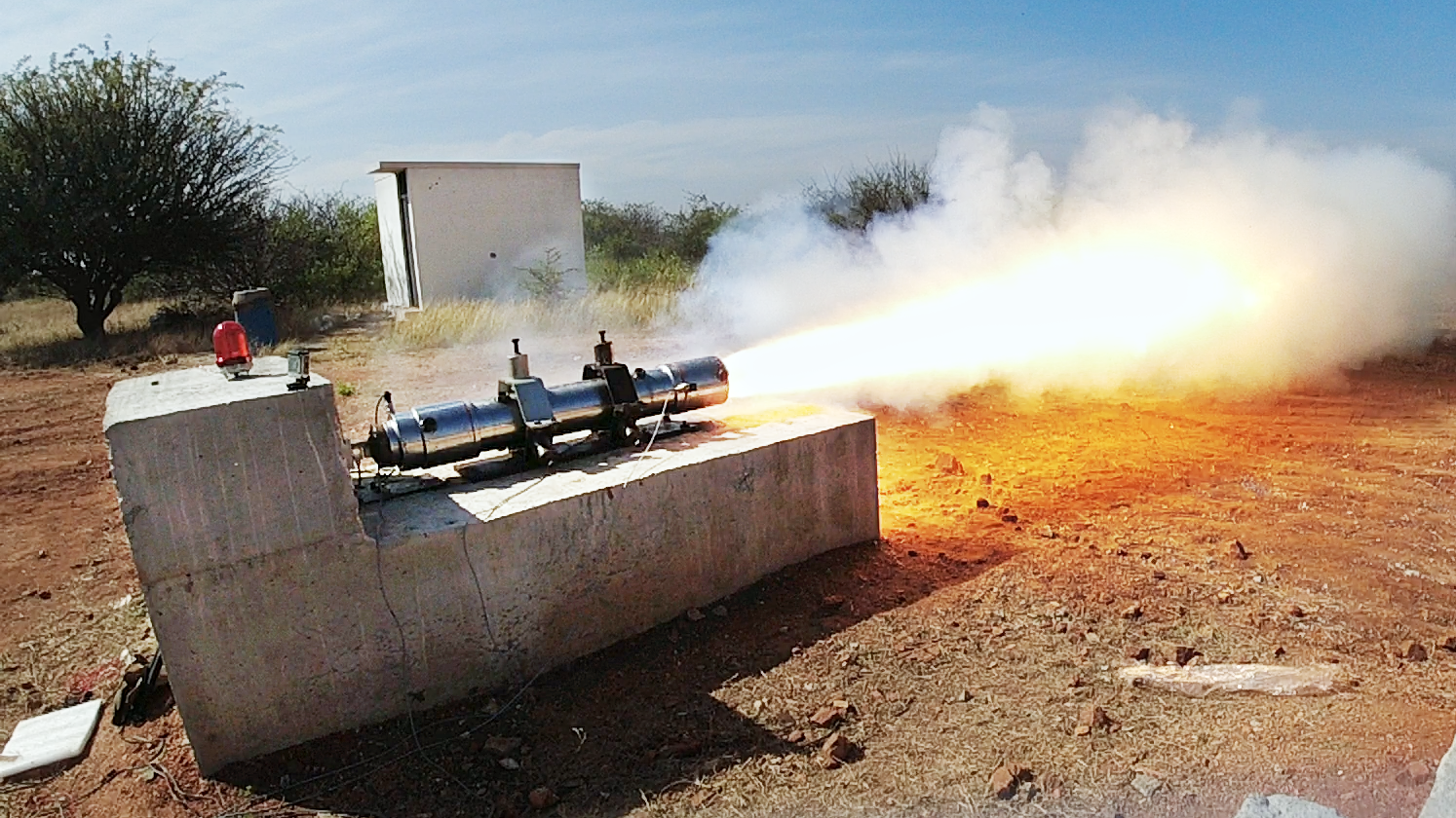 Rocket engine test firing with bright flame and smoke plume from a device on a concrete block.