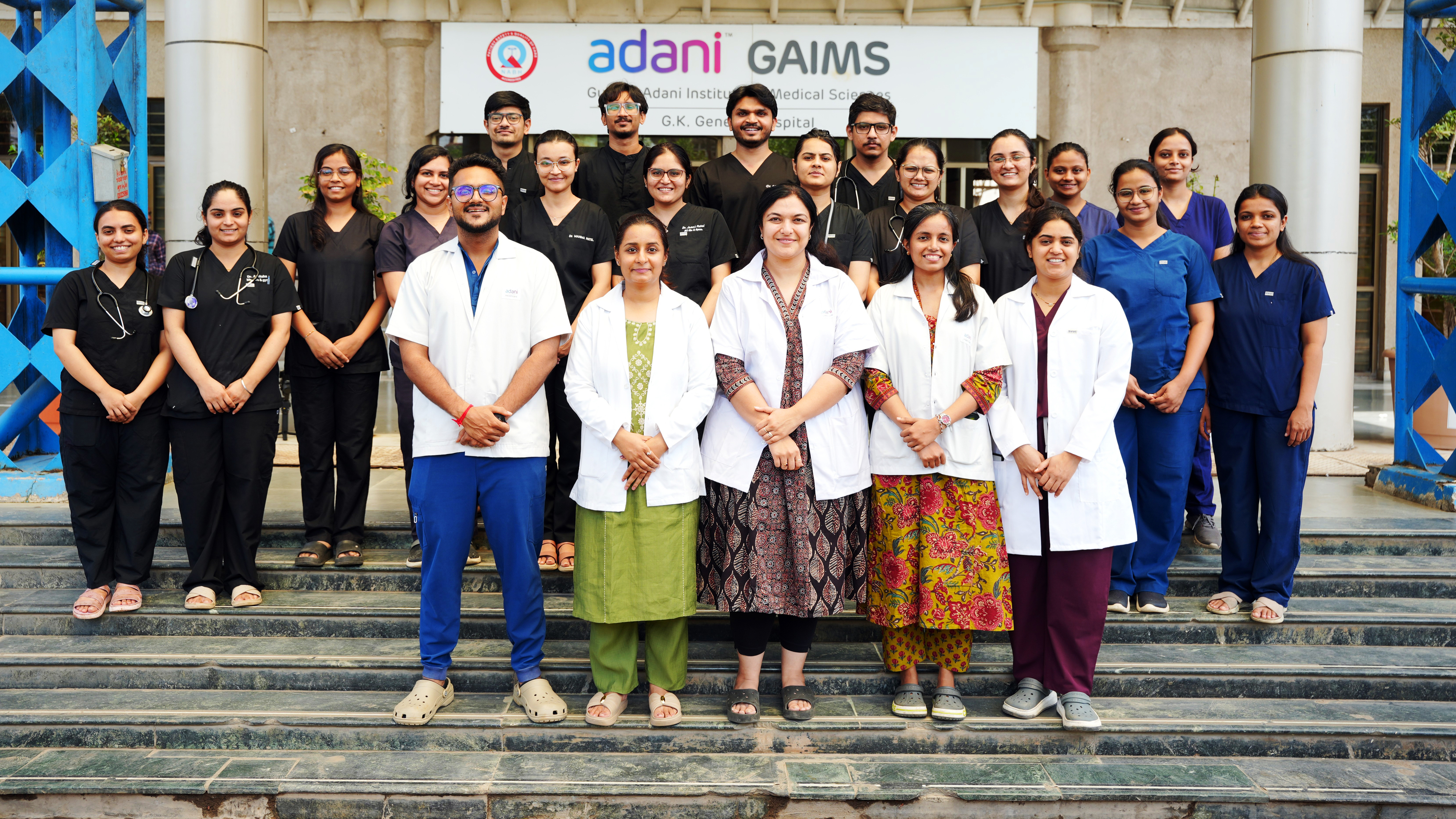A large group of medical professionals in scrubs and lab coats standing on steps in front of Adani GAIMS.