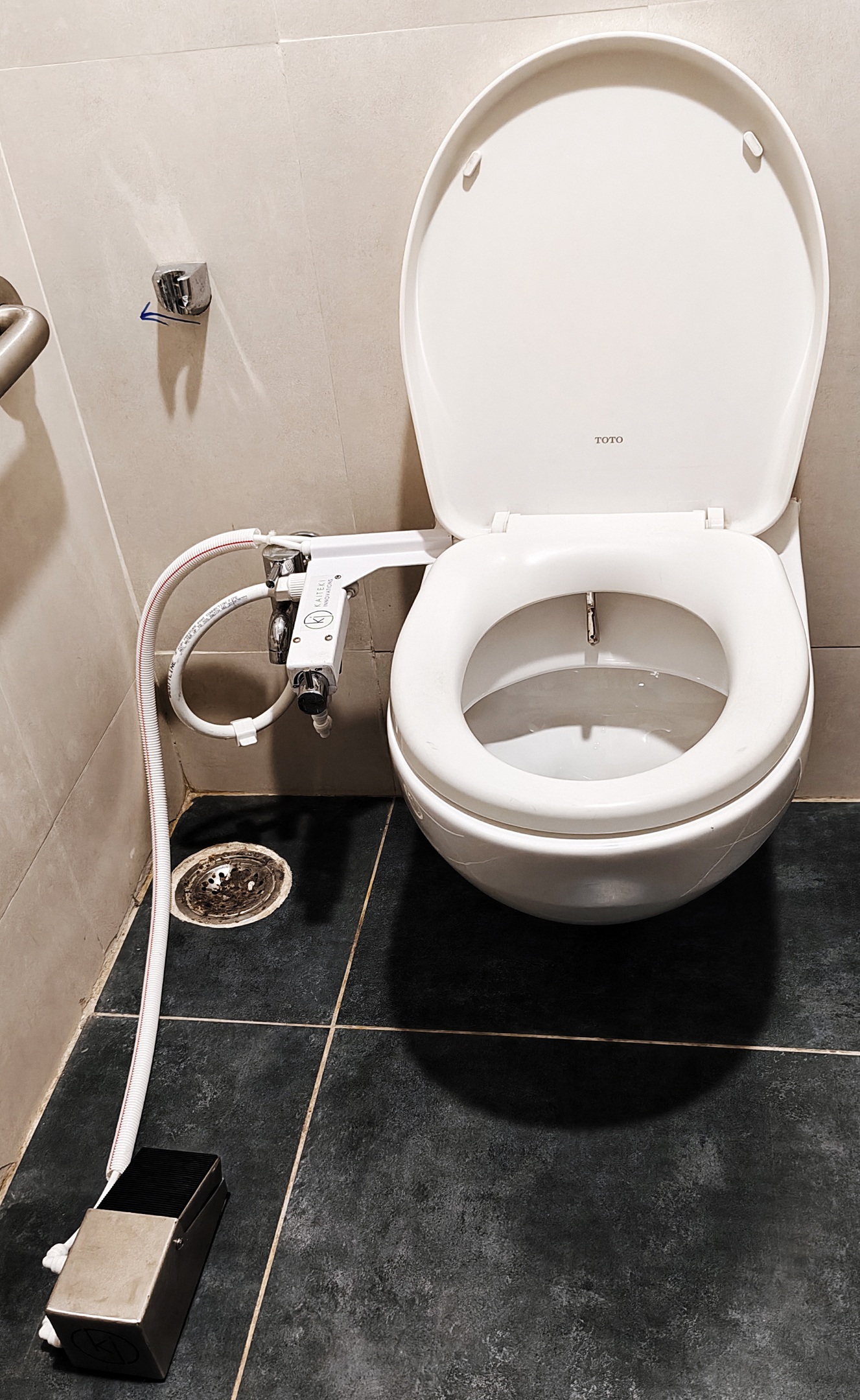 A toilet with a white bidet attachment and a foot pedal on the dark tiled floor.