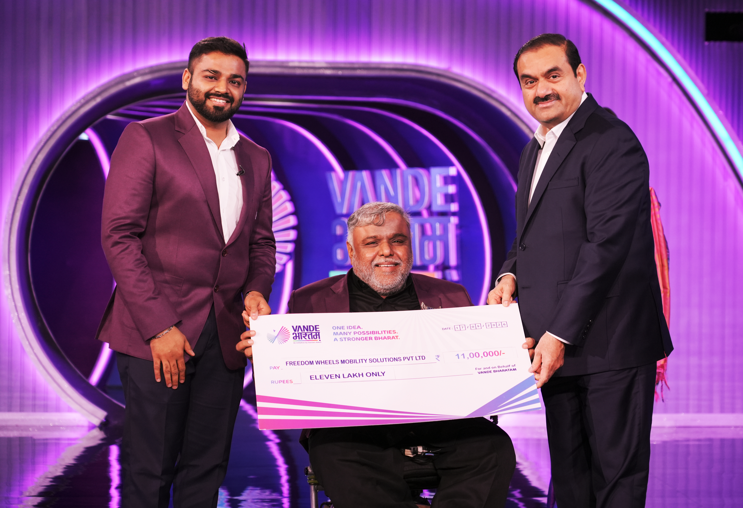 Three men, two standing and one in a wheelchair, hold a large check on a purple stage.