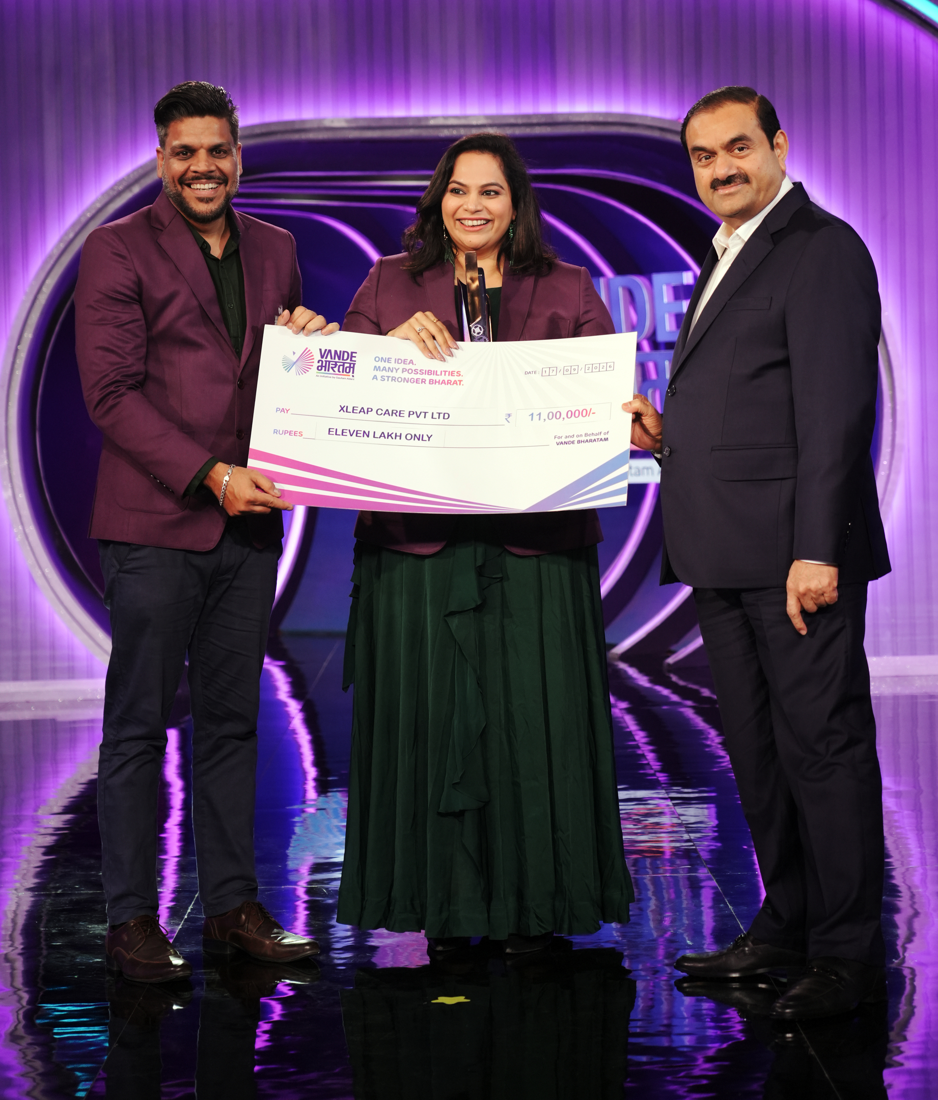 Three people, including Gautam Adani, hold a large check for "Eleven Lakh Only" on a stage.