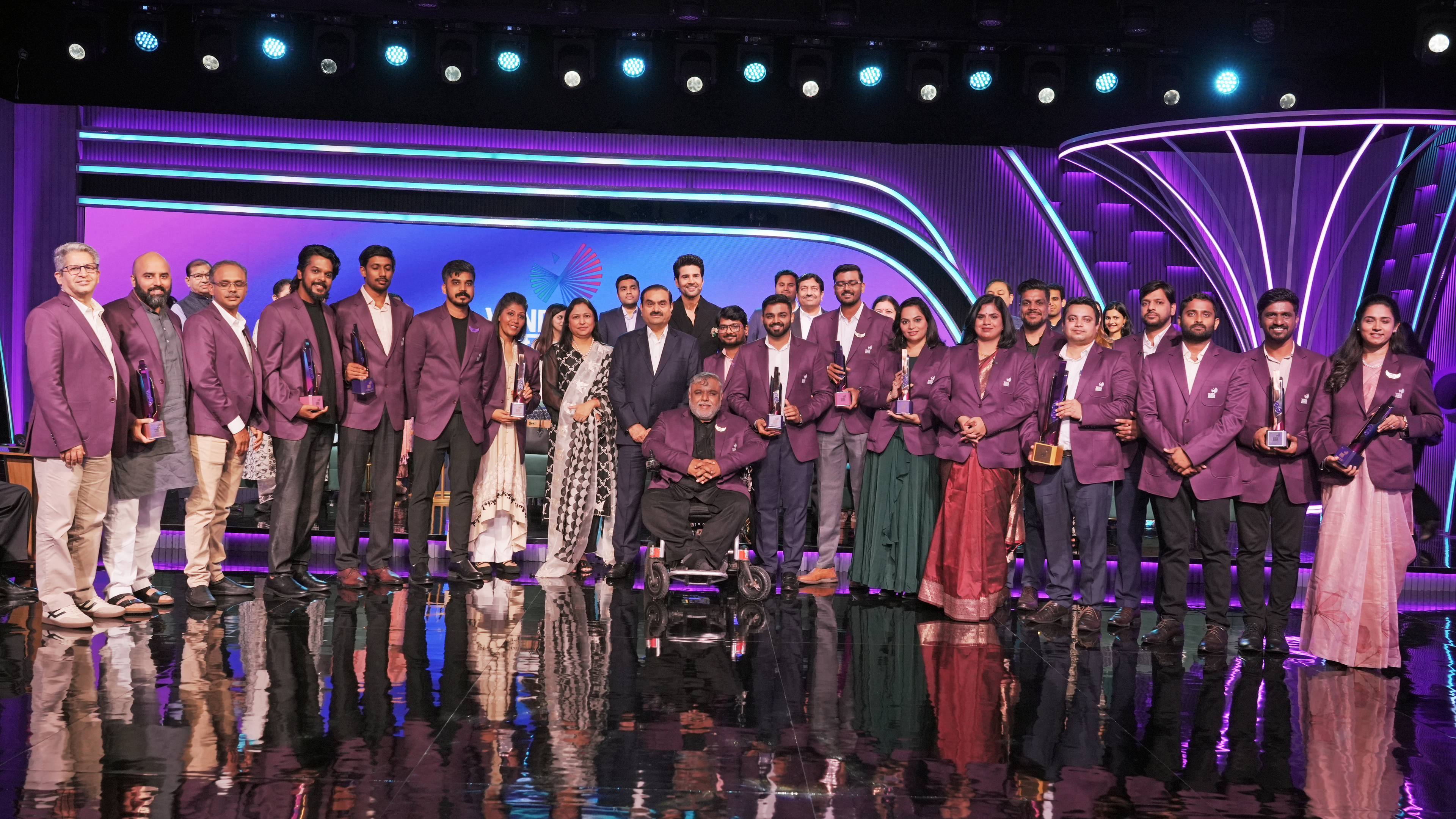 A large group, including Gautam Adani, stands on a stage with awards, purple lighting, and reflections.