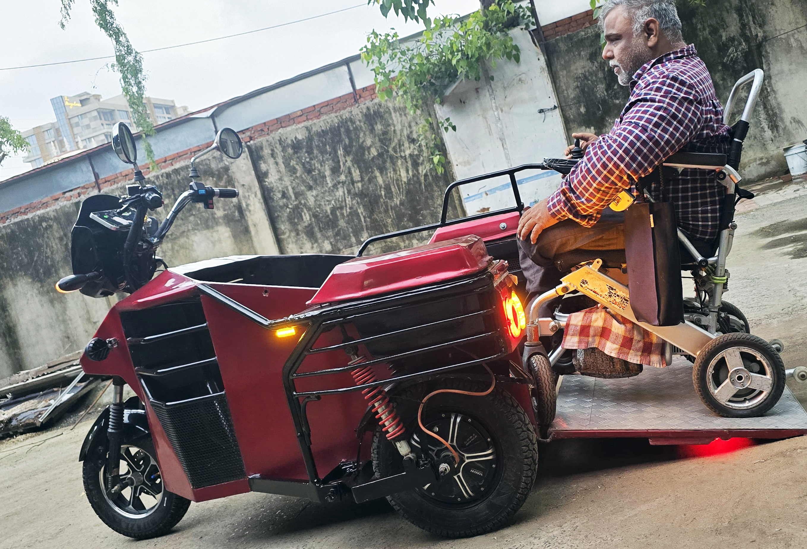 Man in a wheelchair preparing to ride a custom red motorized tricycle with a sidecar.