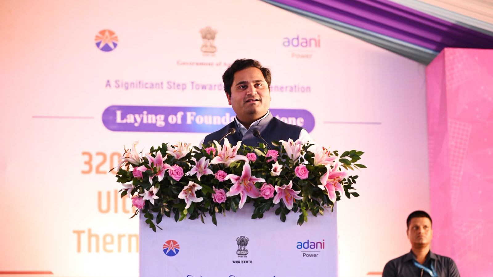 A man in a dark waistcoat speaks at a podium adorned with pink and white flowers during a foundation laying ceremony.