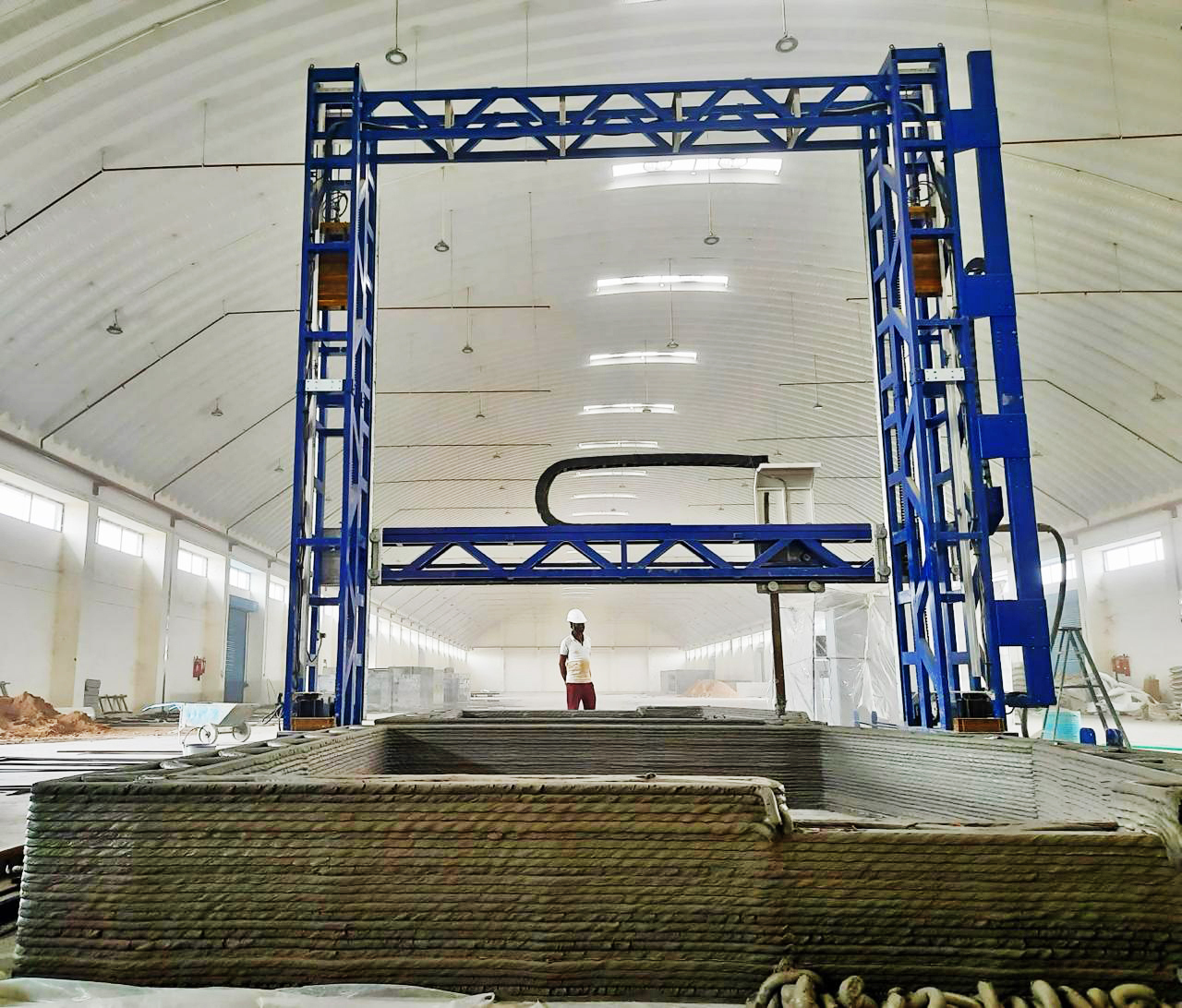 A large blue gantry 3D printer building a concrete structure inside a spacious warehouse, with a worker observing.